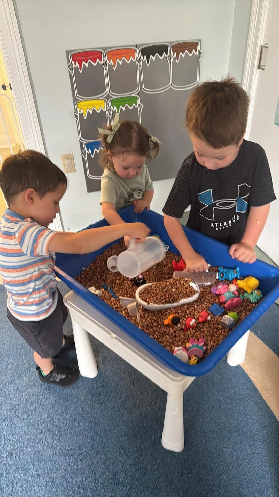 Three children playing with toys and figurines in a sensory table filled with brown coffee beans.