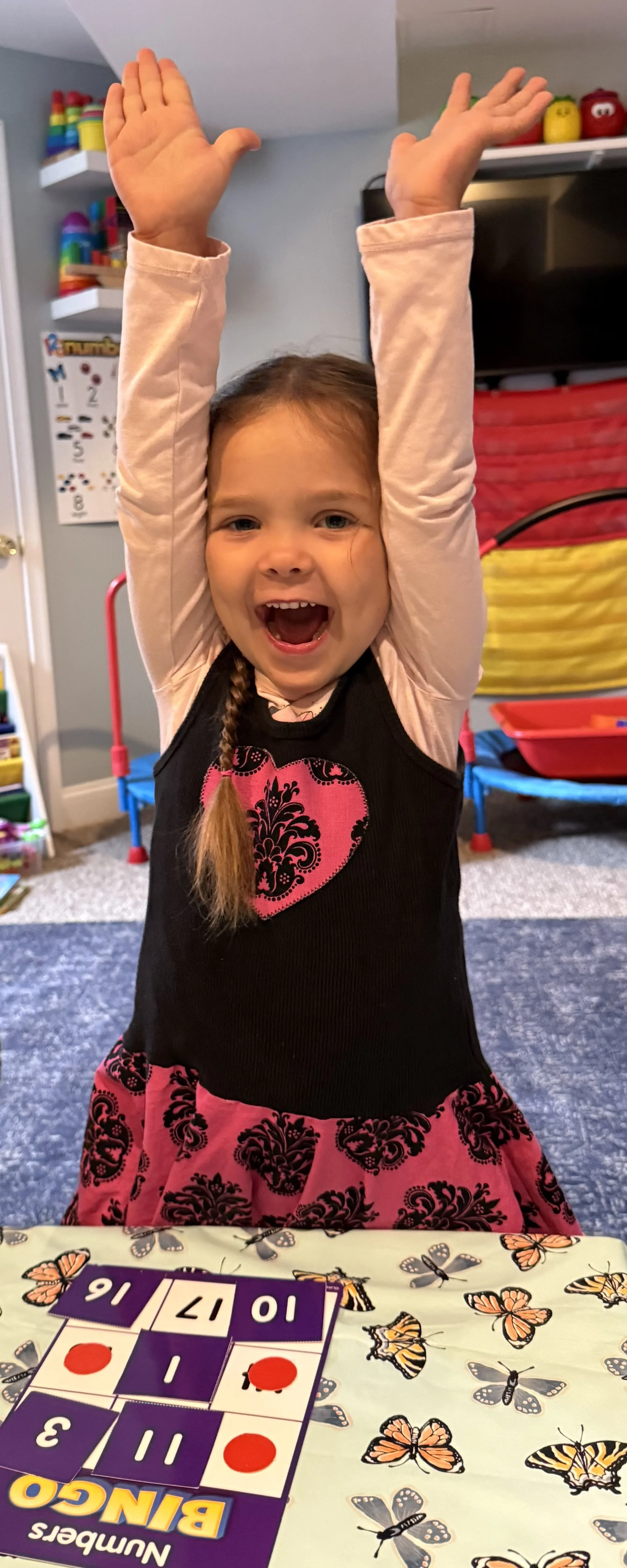 A young girl with light brown hair in a braid, smiling and raising her hands above her head. She is wearing a black dress with pink and black heart and floral patterns. She is sitting at a table with a butterfly-themed tablecloth, playing a game with