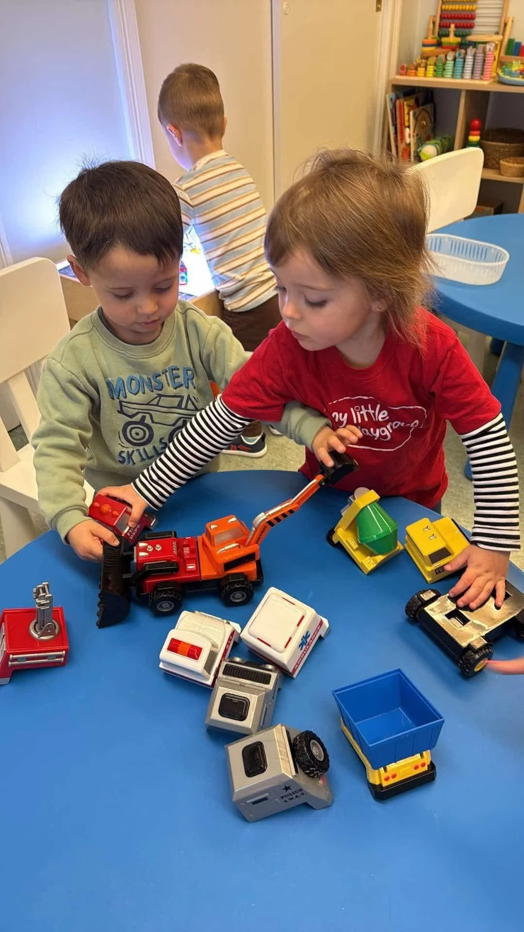 Two children playing with toy rescue vehicles at a blue table, with one boy in a gray shirt and girl in a red shirt.
