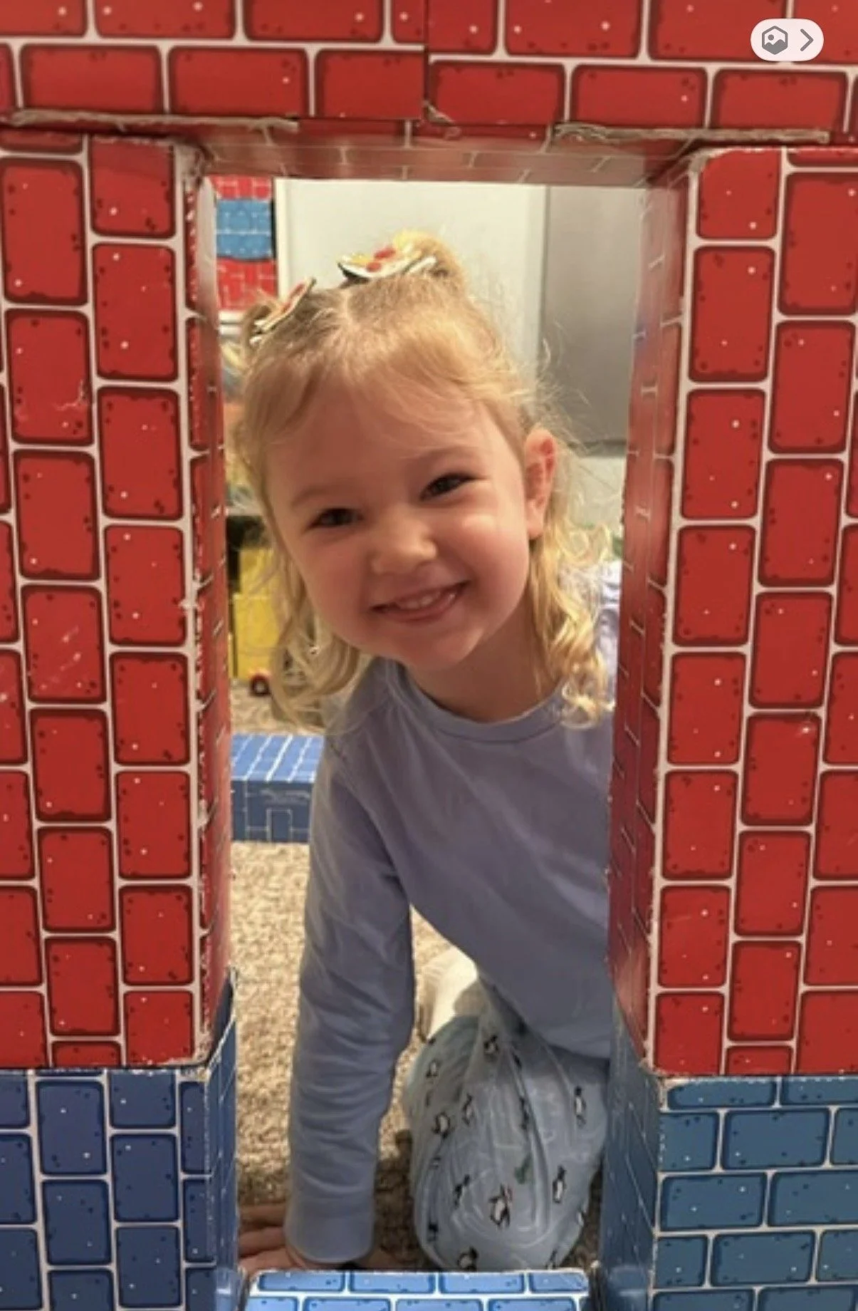 A young girl with blonde hair and a bow in her hair, smiling through a cardboard structure decorated to look like a brick wall, with red and blue bricks, in a playroom.