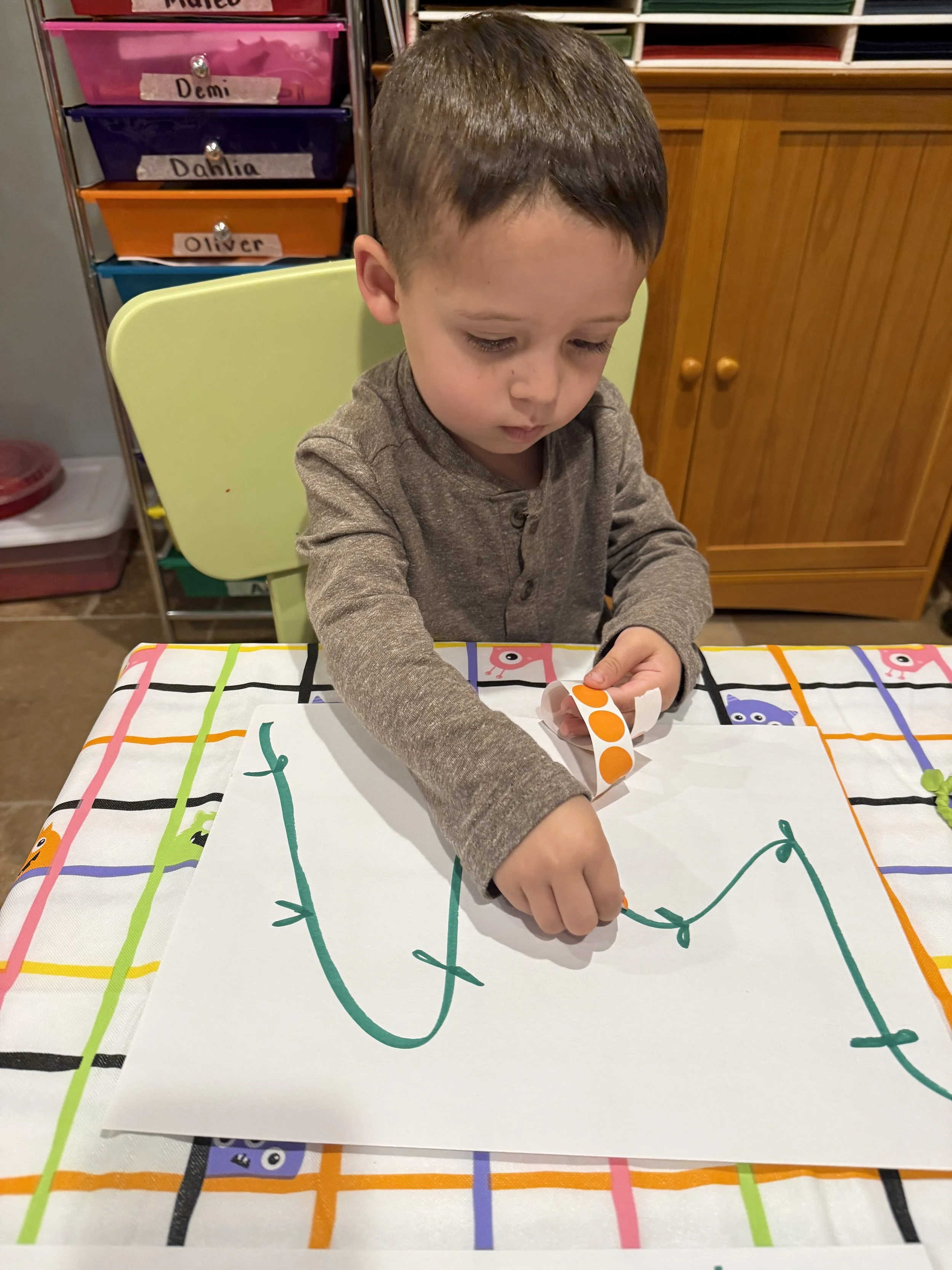 A young boy sitting at a table, drawing with a green marker on a large white sheet of paper with colorful grid lines and animal illustrations.