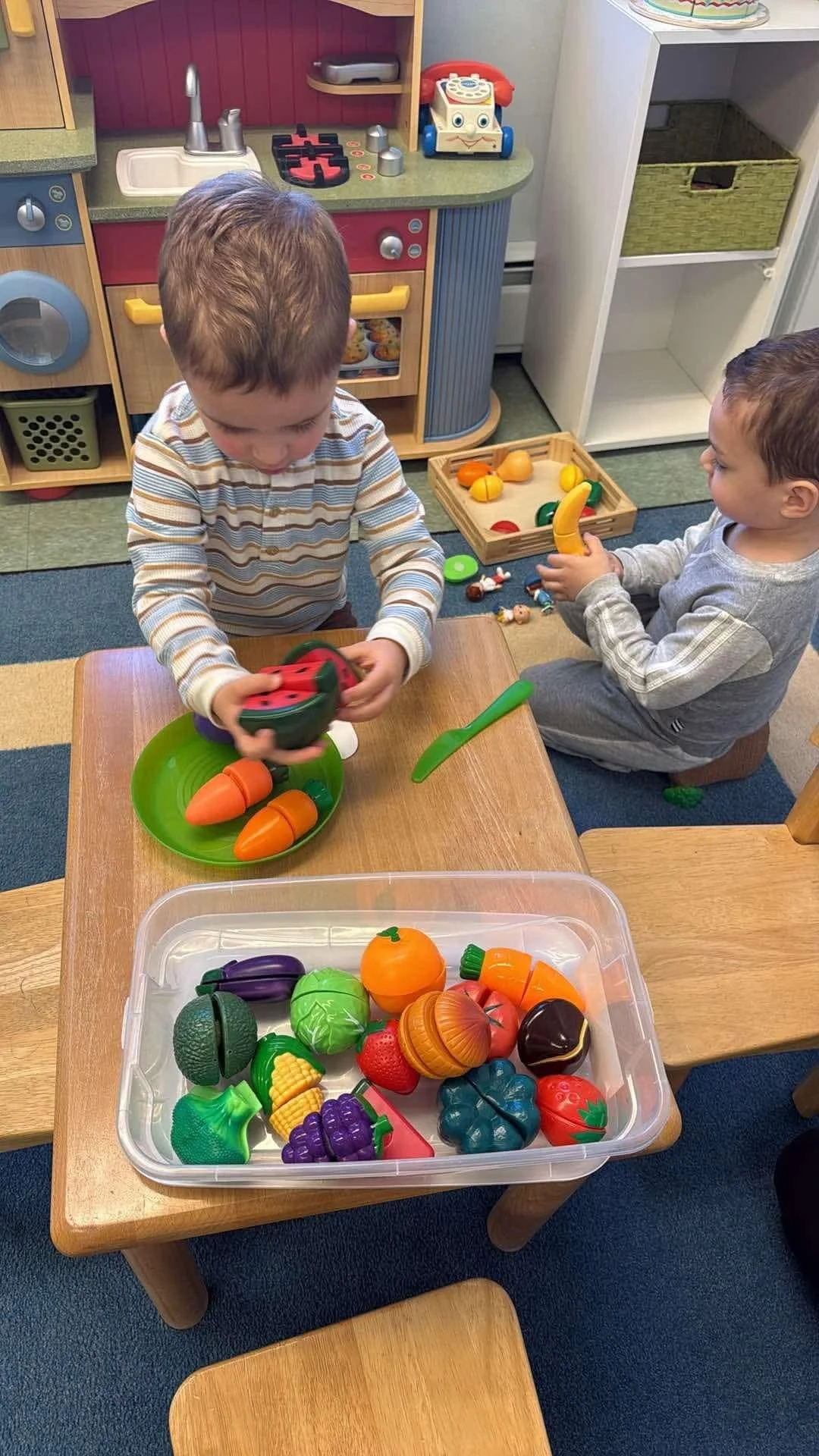 Two young boys playing with toy food and grocery items in a play kitchen space. One boy is holding a toy watermelon, and the other boy is holding a toy banana.