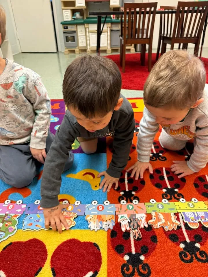 Three young children playing on a colorful rug with number and animal-themed puzzle pieces. The children are kneeling and focusing on fitting the pieces together.