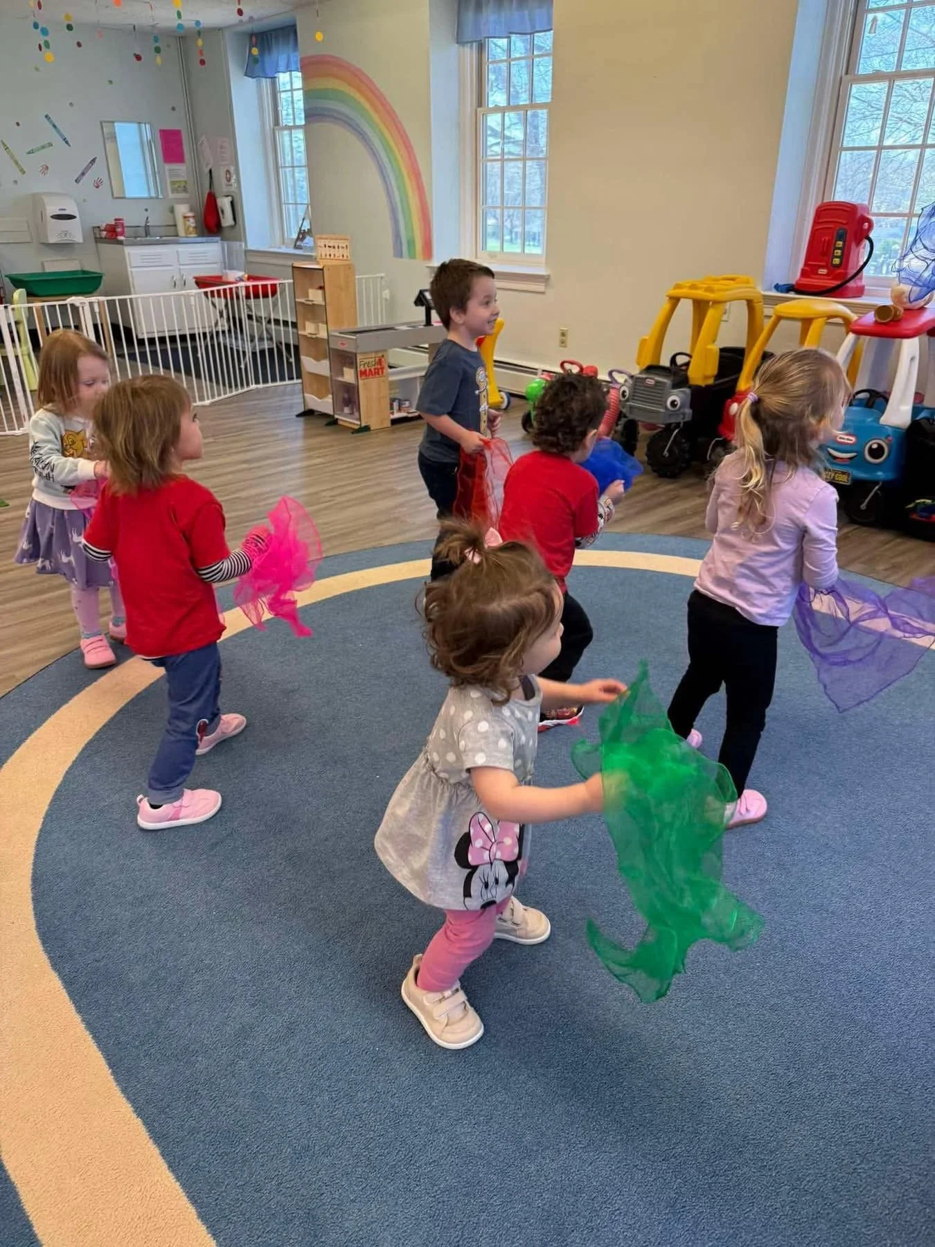 Children playing with colorful scarves in a classroom with toy riding cars and a rainbow painted on the wall.