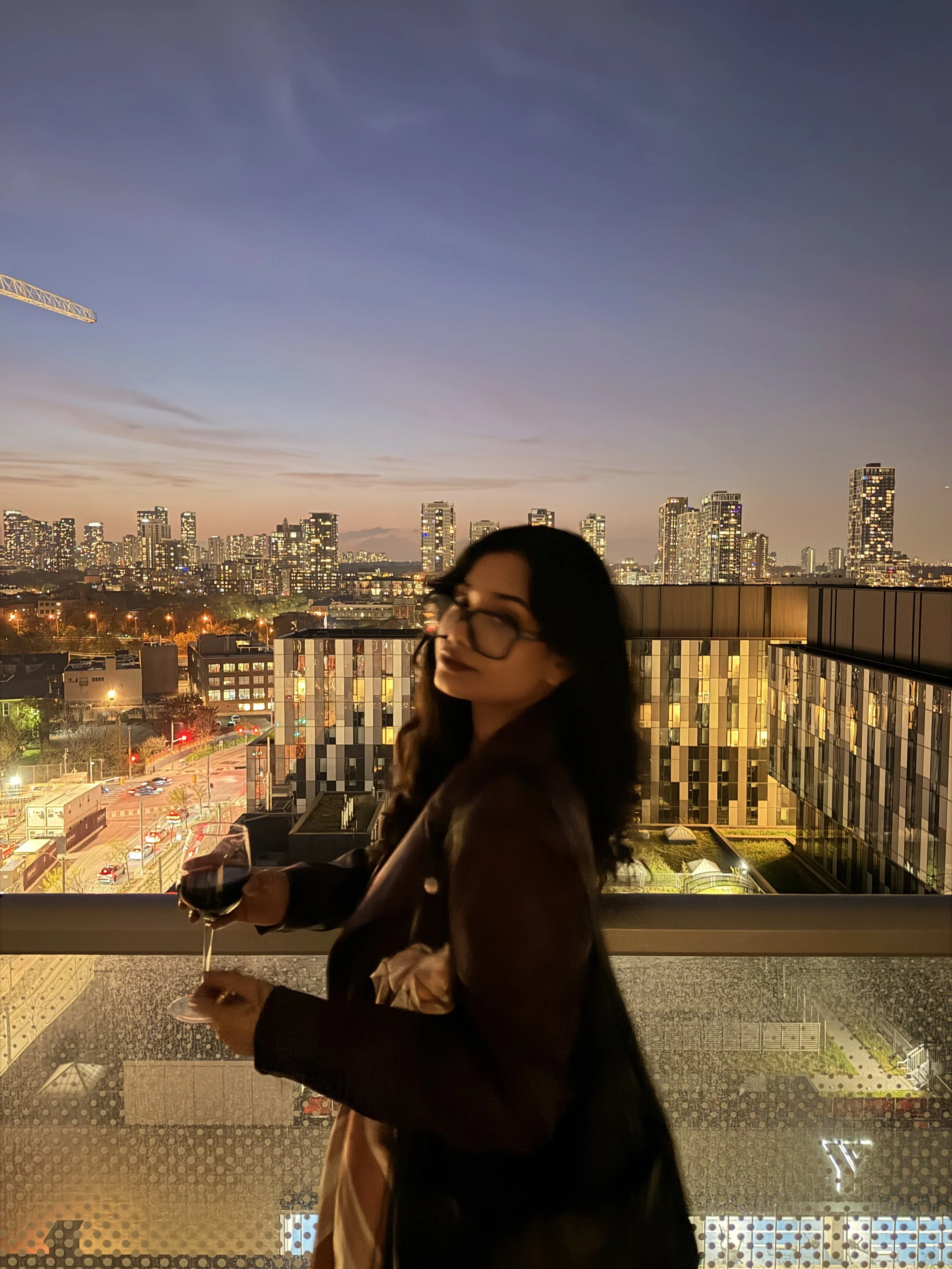 A woman holding a glass of red wine stands on a balcony overlooking a city skyline at dusk or nighttime. The city buildings are lit up, and the sky shows a gradient from sunset hues to dark blue.