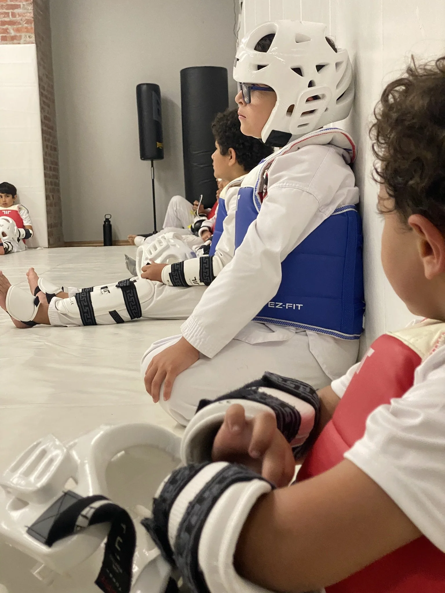 Group of children sitting on the floor wearing martial arts uniforms and gear, including helmets, with an indoor setting featuring a brick wall and speakers in the background.
