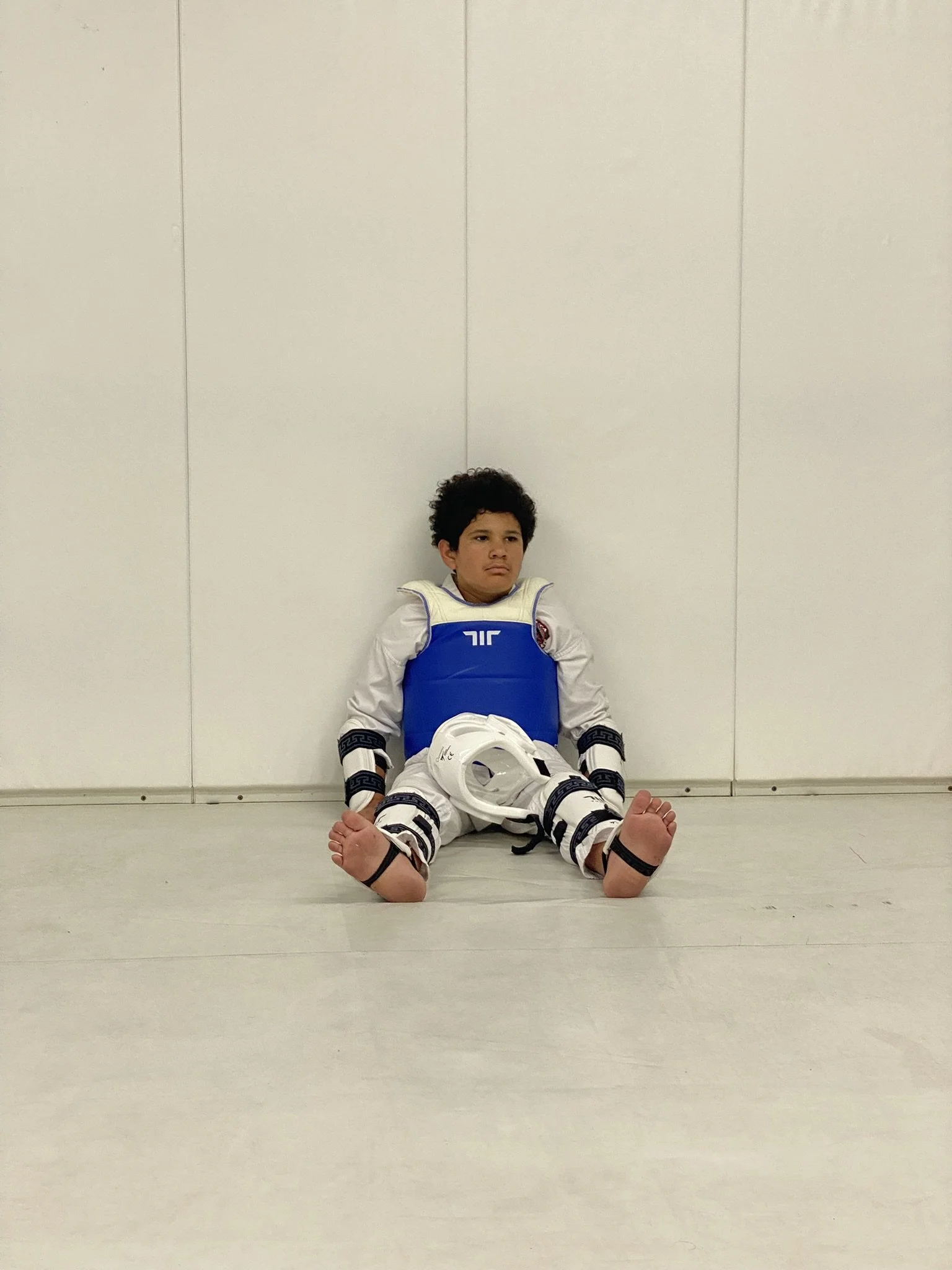 Young boy sitting on the floor in a martial arts uniform with a blue chest protector, white helmet, and black wristbands, against a plain white wall.