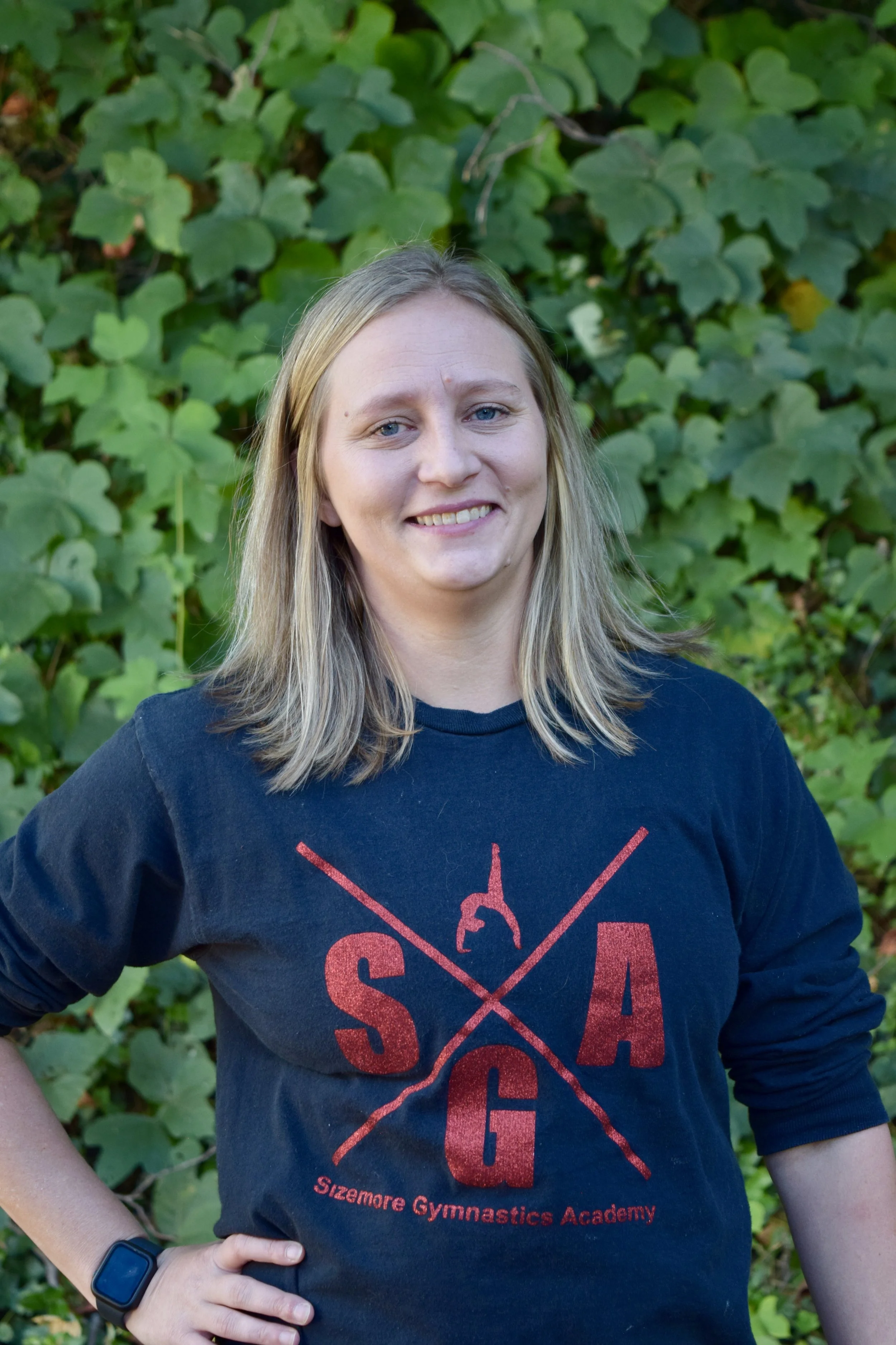 A woman with blonde hair wearing a black T-shirt with red text and graphic, standing outdoors in front of green foliage.