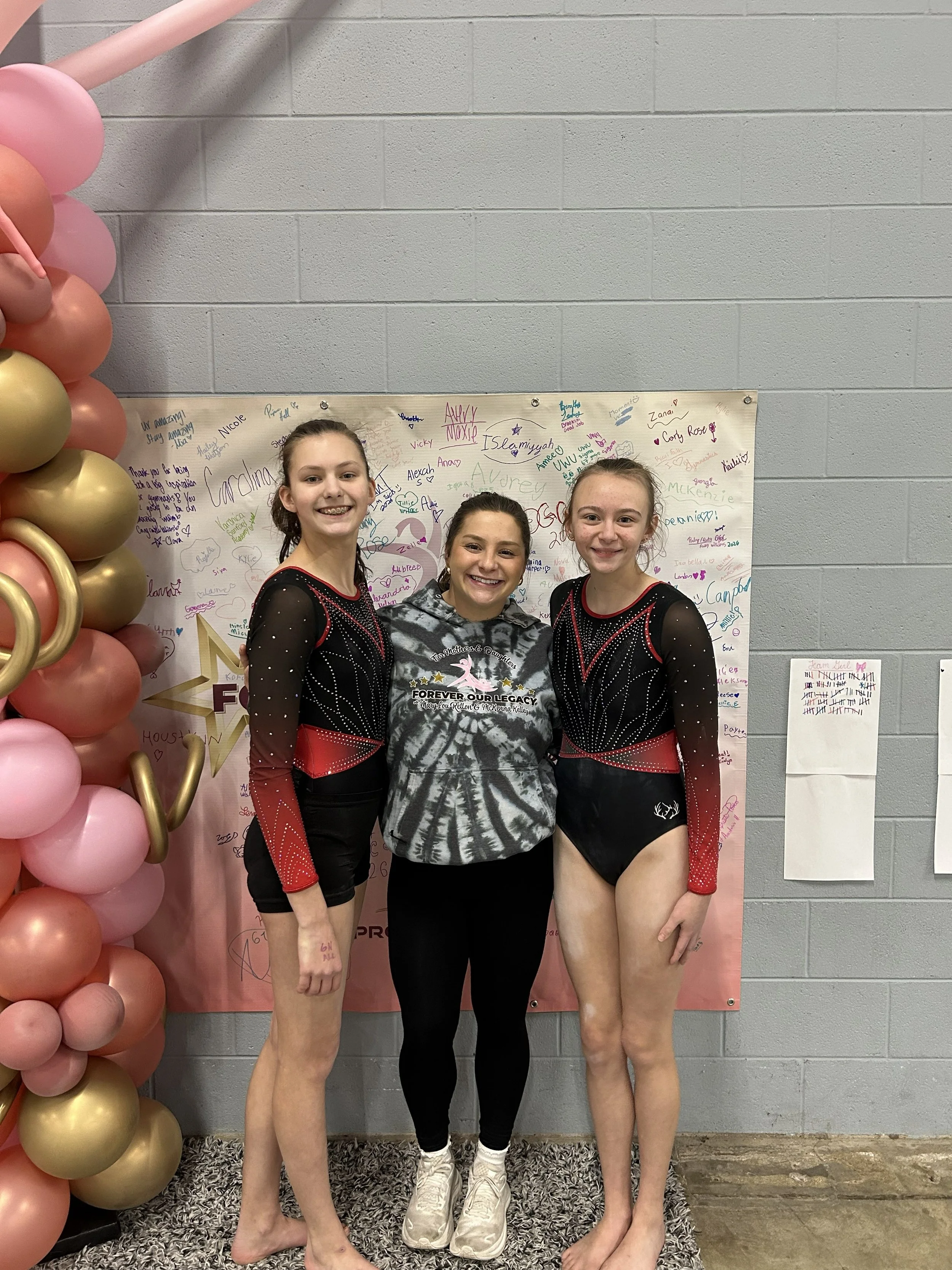 Three young female gymnasts in black and red costumes standing next to a woman in athletic wear, posing in front of a decorated backdrop with balloon arrangements and signatures, at an indoor event.