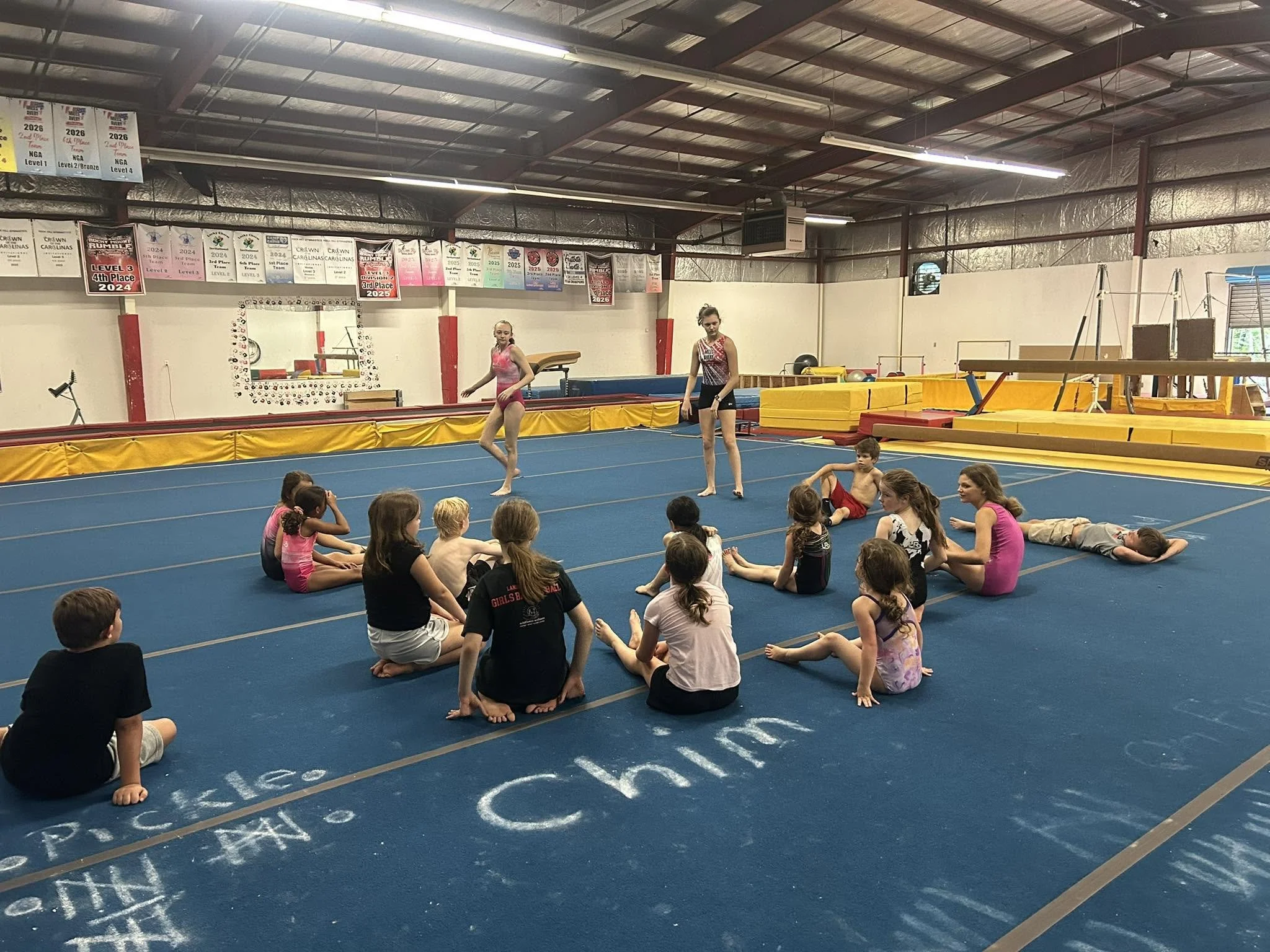 Children sitting on a blue gym floor watching two girls practice gymnastics. The gym has banners hanging on the wall celebrating gymnastics achievements.