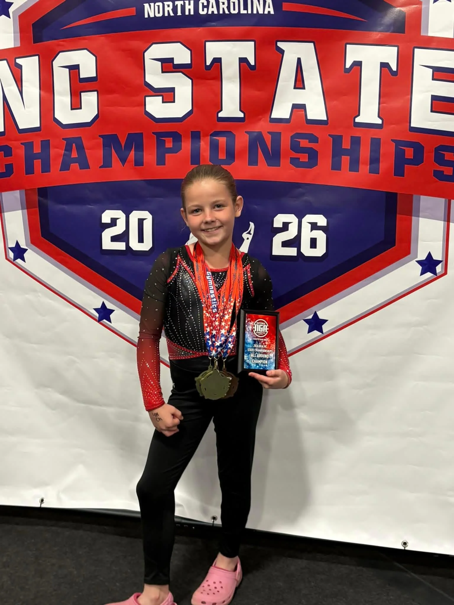 Young girl smiling, standing in front of a North Carolina gymnastics championship banner, holding a plaque and wearing medals around her neck.
