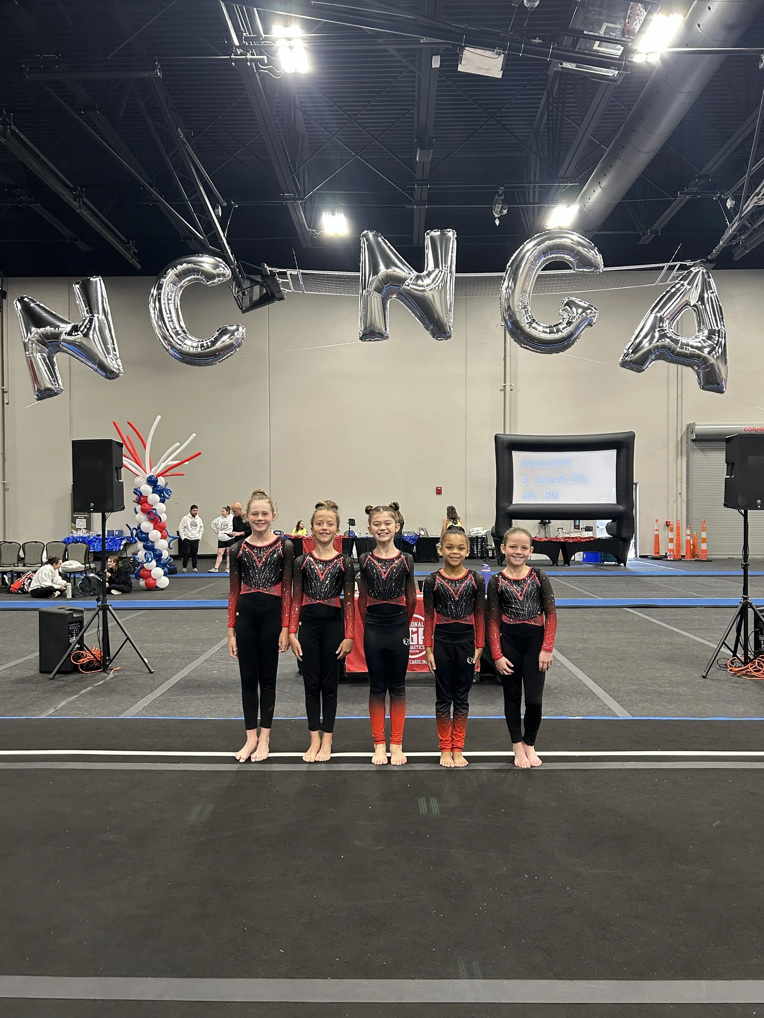 Five young female gymnasts stand in a line on a competition floor, smiling at the camera, showcasing their red and black leotards. Behind them, large silver balloon letters spell out 'NCGA,' hanging from the ceiling, with some balloon decorations and event setup visible in the background.