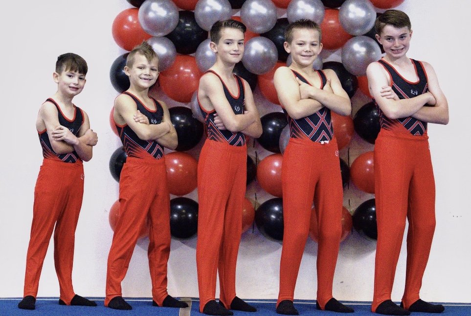 Five young boys in matching gymnastics outfits standing with arms crossed in front of a backdrop of balloons in red, black, and silver.