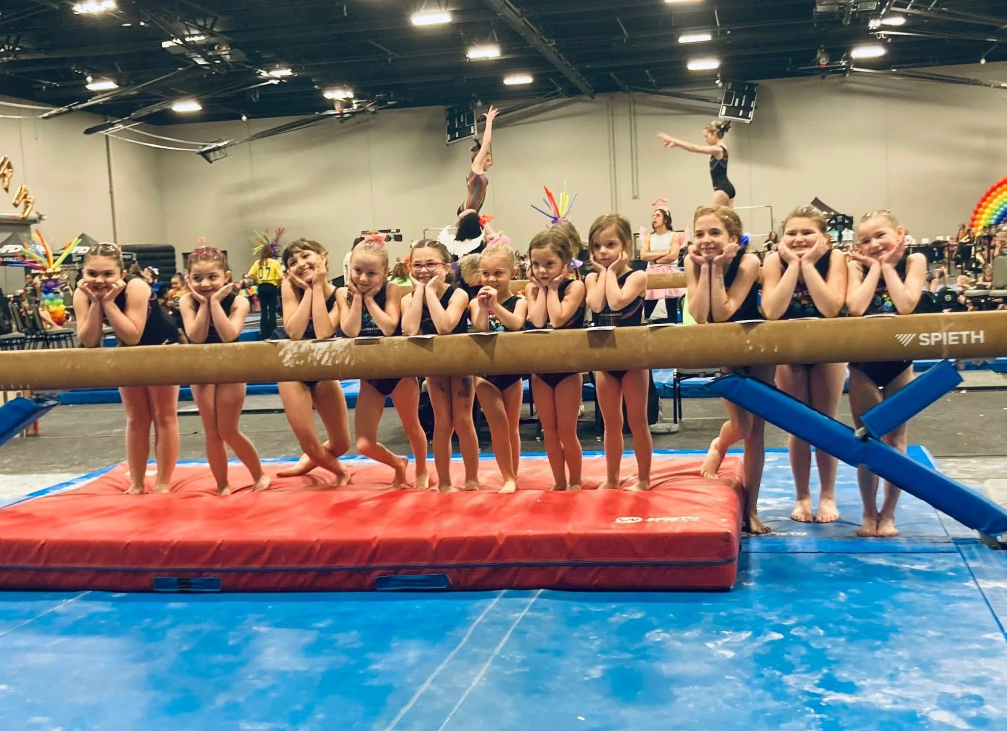 Group of young girls sitting behind a balance beam during a gymnastics meet, with two girls performing flips in the background and decorated for the event.