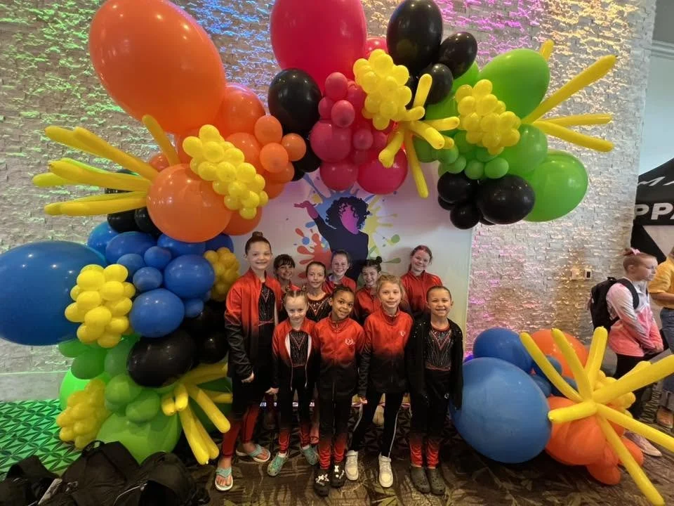 Group of young girls in matching black and red outfits posing in front of a colorful balloon arch at an event.