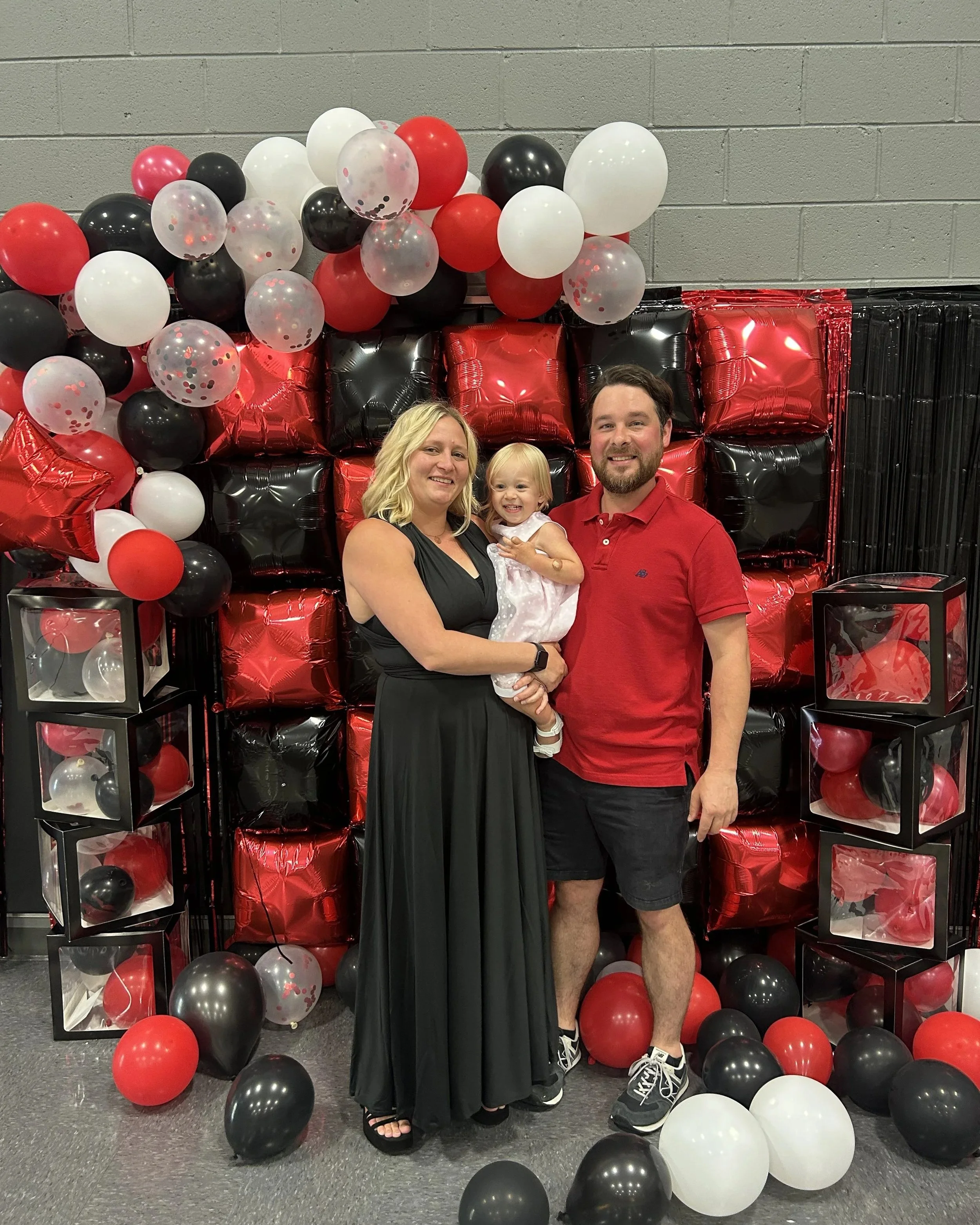 A family of three, with a woman, a man, and a young girl, standing in front of a backdrop made of red, black, and white balloons, and smiling. The woman has blonde hair and wears a black dress, the man has a beard, wears a red polo shirt and shorts, and the girl has blonde hair, a white dress with pink and purple details, and white shoes.