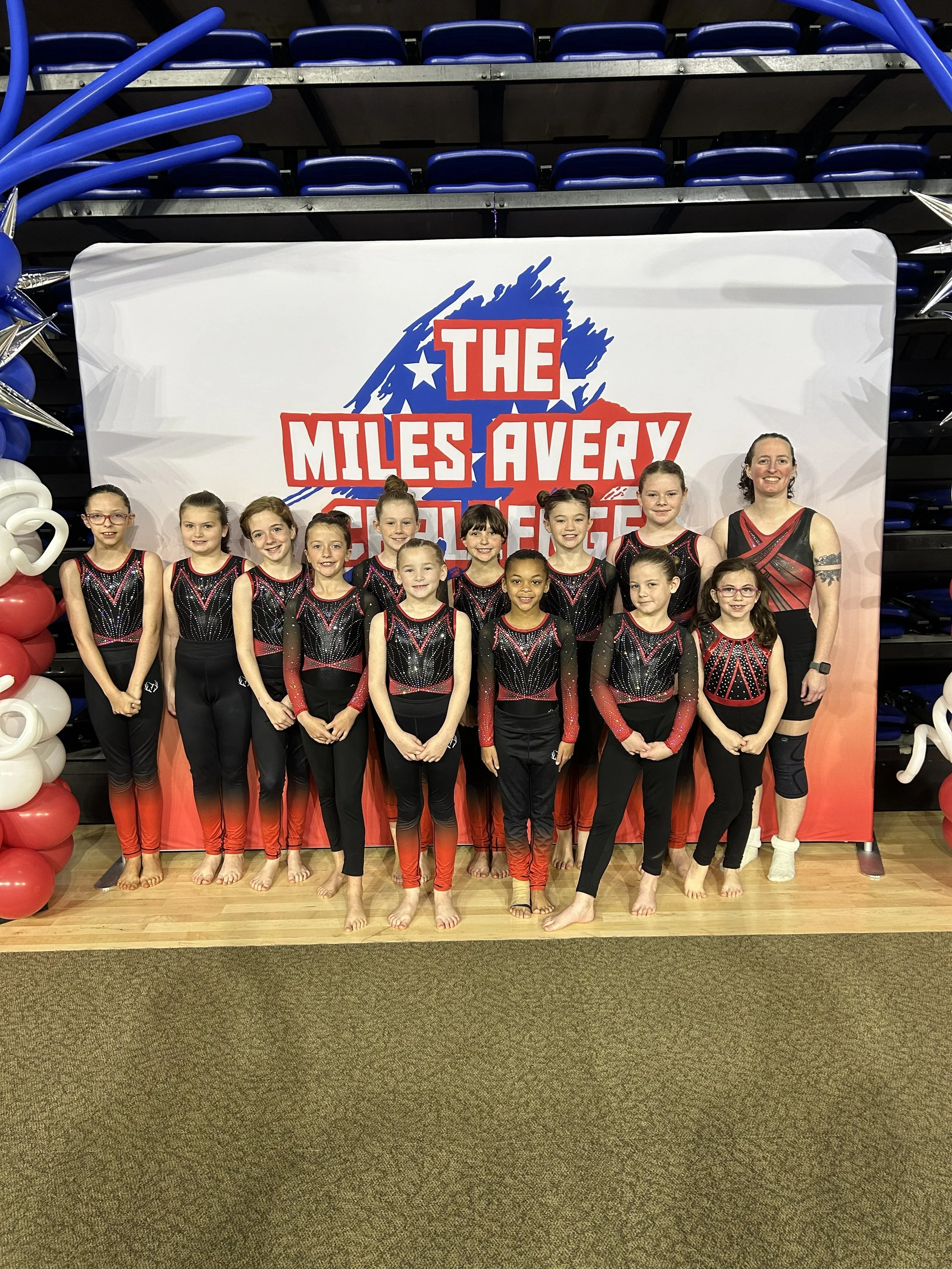 A group of young female gymnasts in black and red leotards, standing in front of a backdrop that reads "The Miles Avery", inside a gymnasium with blue bleachers and balloons on the sides.