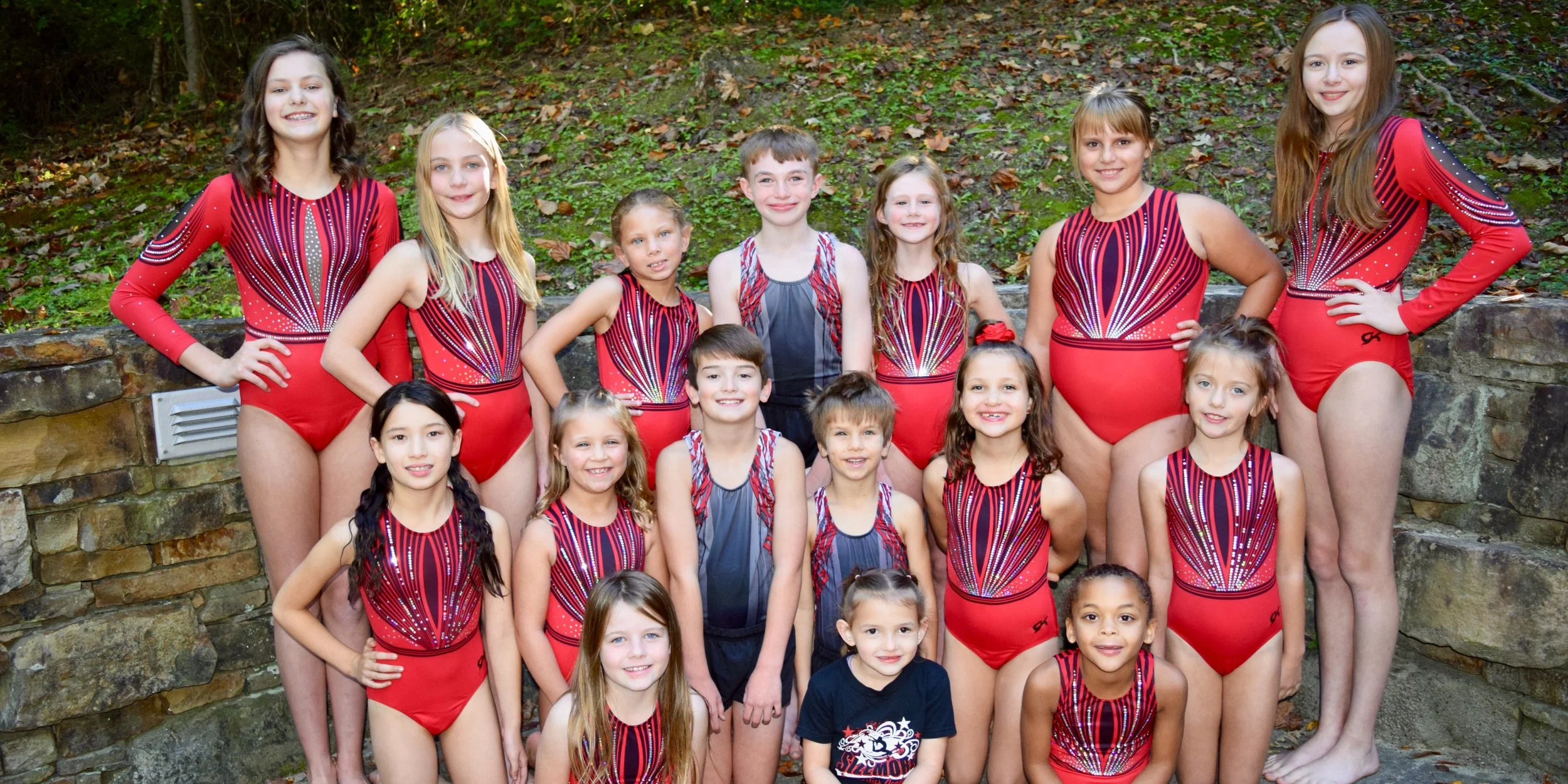 A group of children in red and black gymnastics leotards posing outdoors on a stone ledge with green foliage in the background.