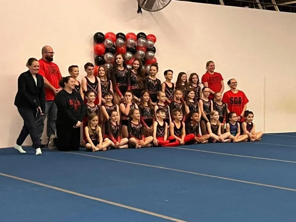 Group of young gymnasts and coaches posing for a photo at a gymnastics event, with black, red, and silver balloons in the background.