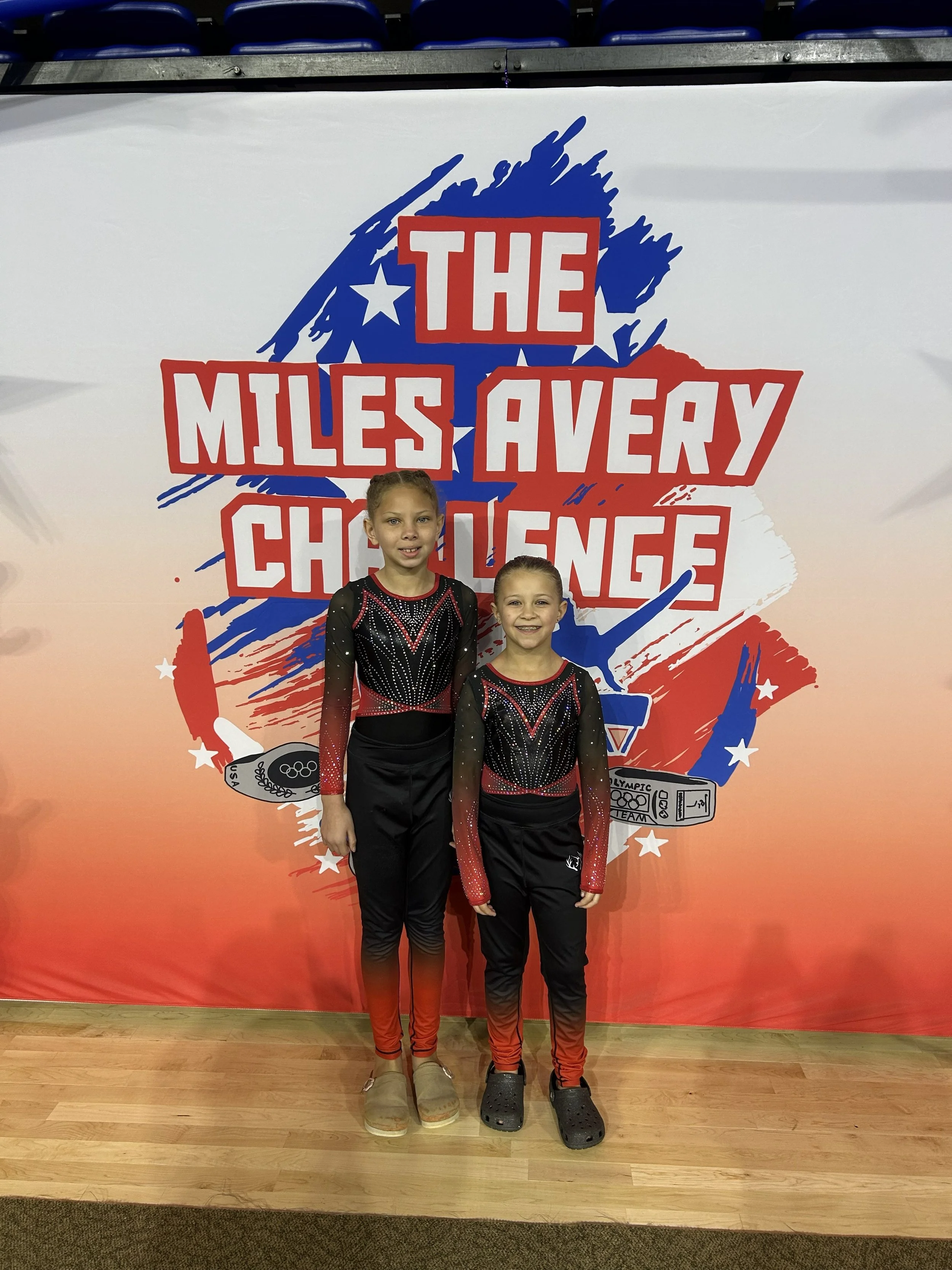 Two young girls dressed in matching black and red cheerleading or gymnastics outfits standing in front of a large event banner that reads 'The Miles Avery Challenge' with stars and an illustration of a medal, on a wooden floor.