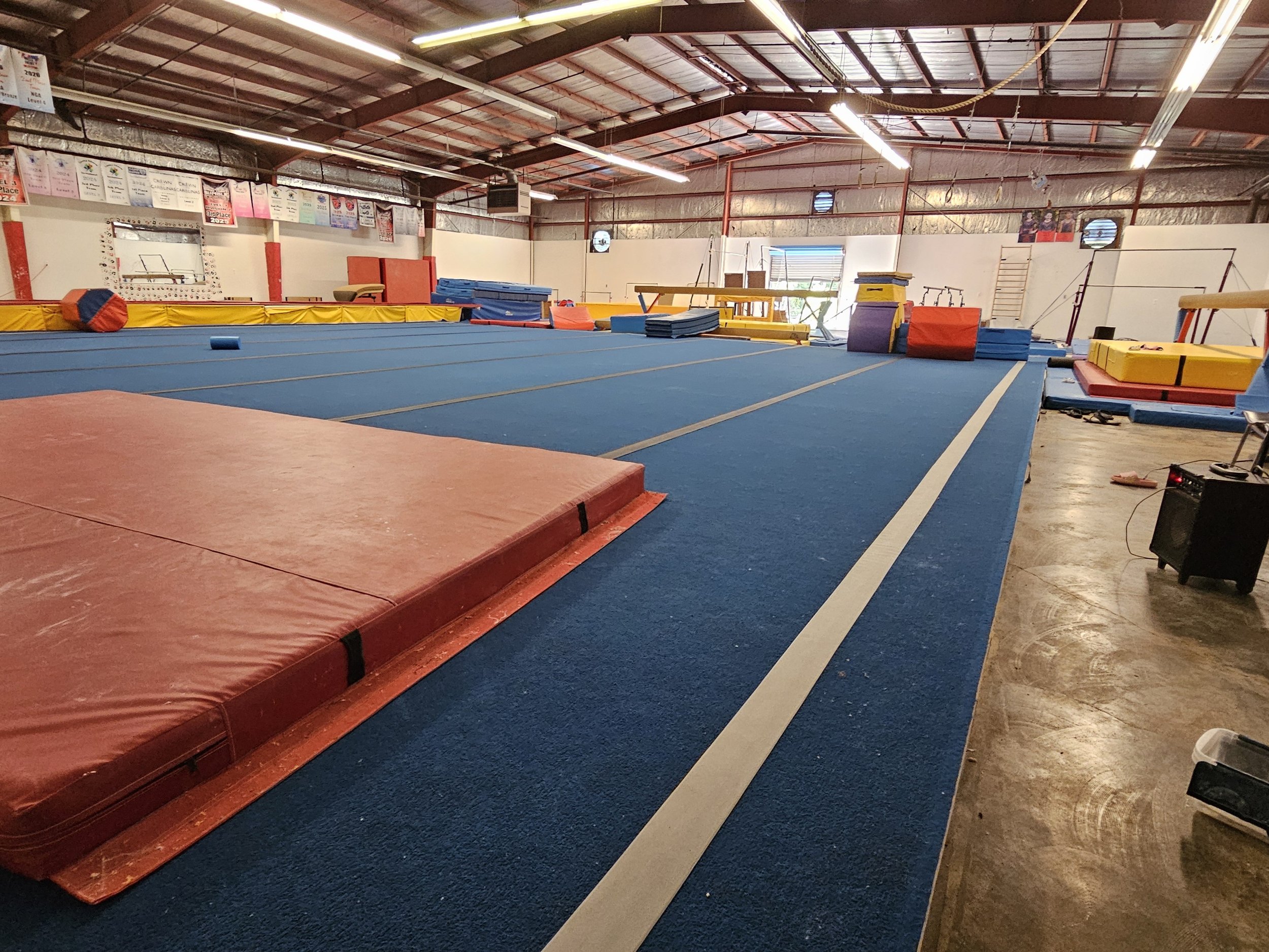 Indoor gymnastics training facility with blue mats, red and yellow padded equipment, and banners hanging from the ceiling.