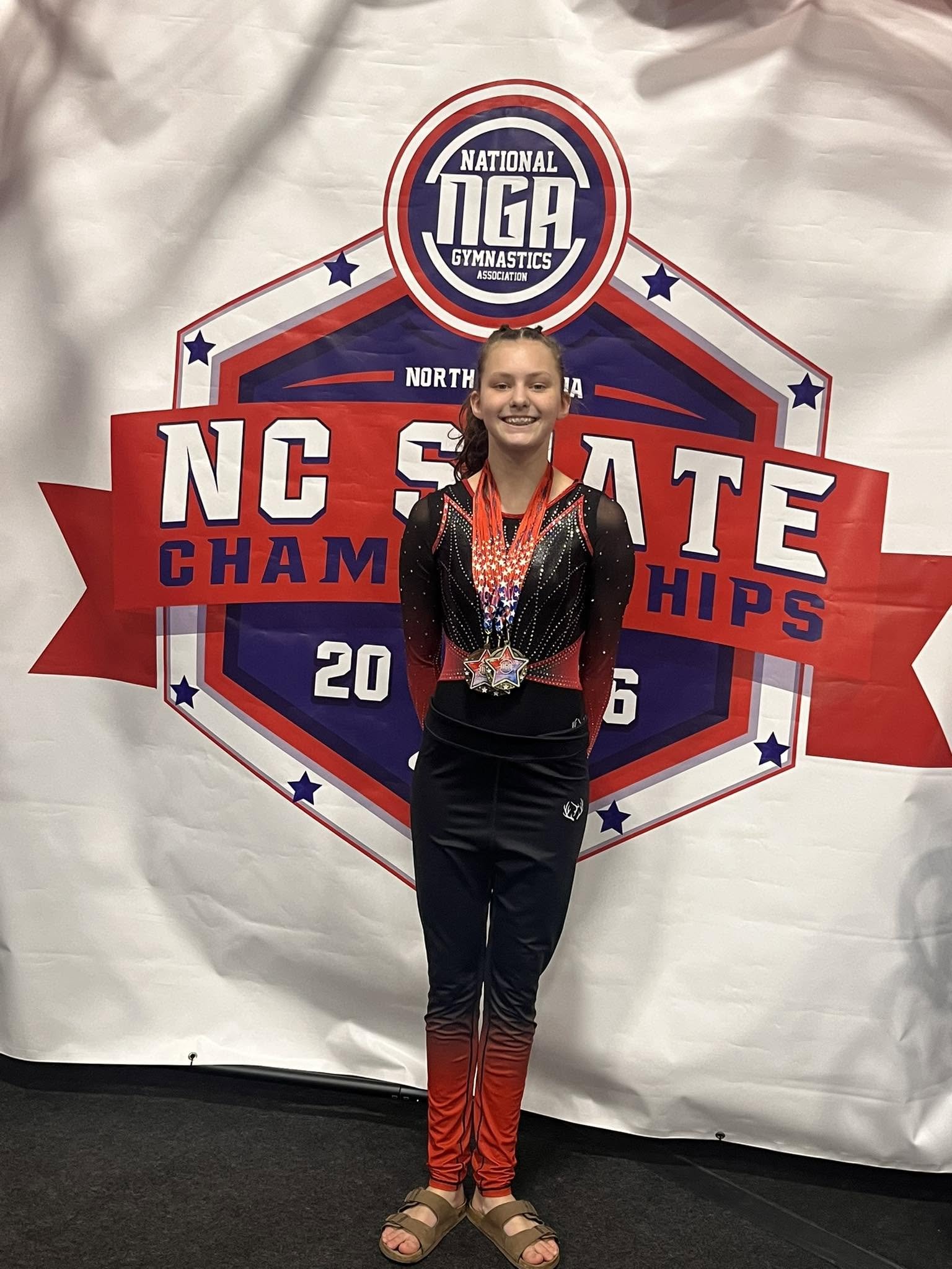 A young girl in gymnastics attire standing in front of a banner celebrating North Carolina State Championships. She is wearing a black and red leotard with rhinestones and a medal around her neck, smiling at the camera.
