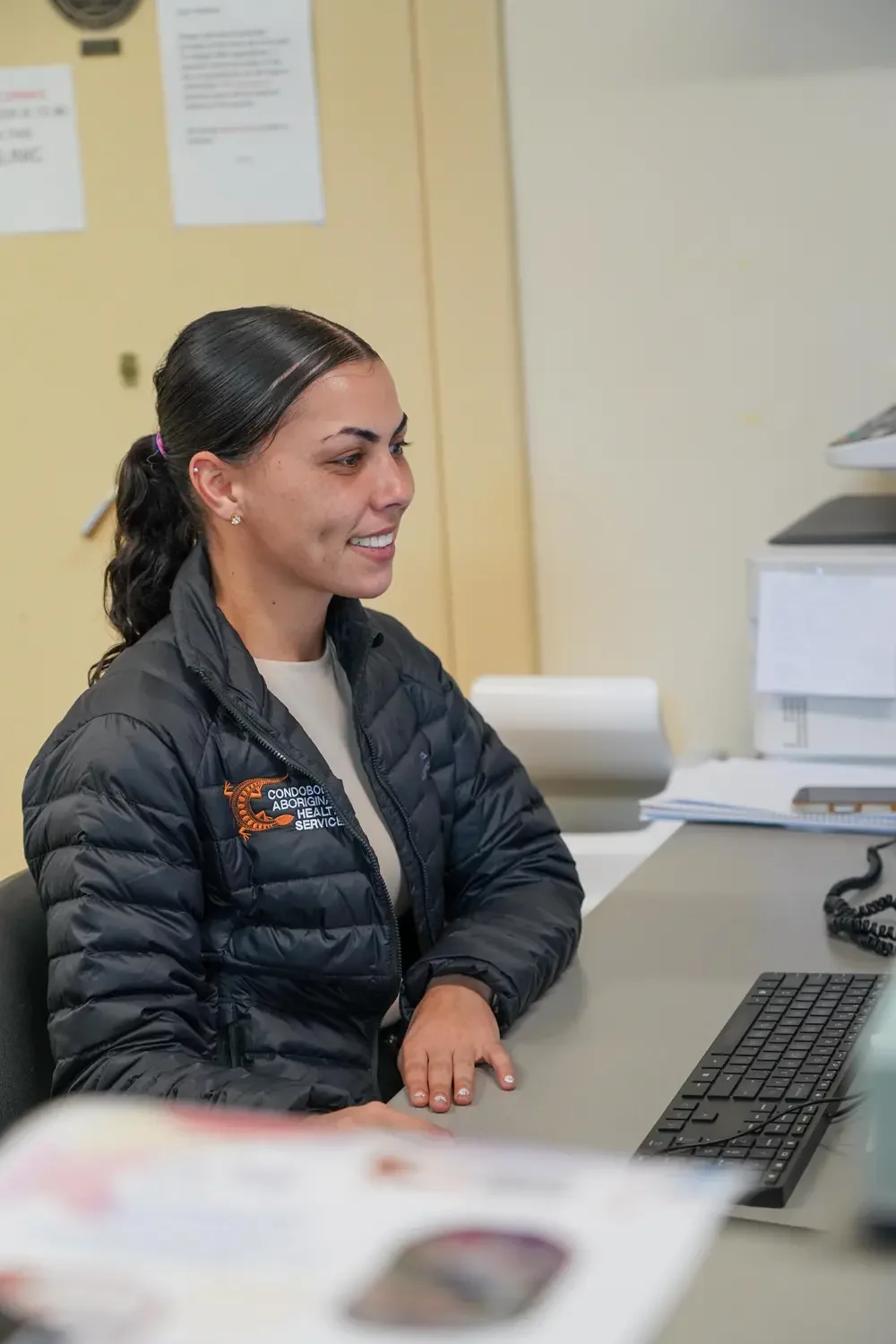 A woman sitting at a desk in an office, wearing a black jacket with a logo that reads 'Condobol Aboriginal Health Service'.