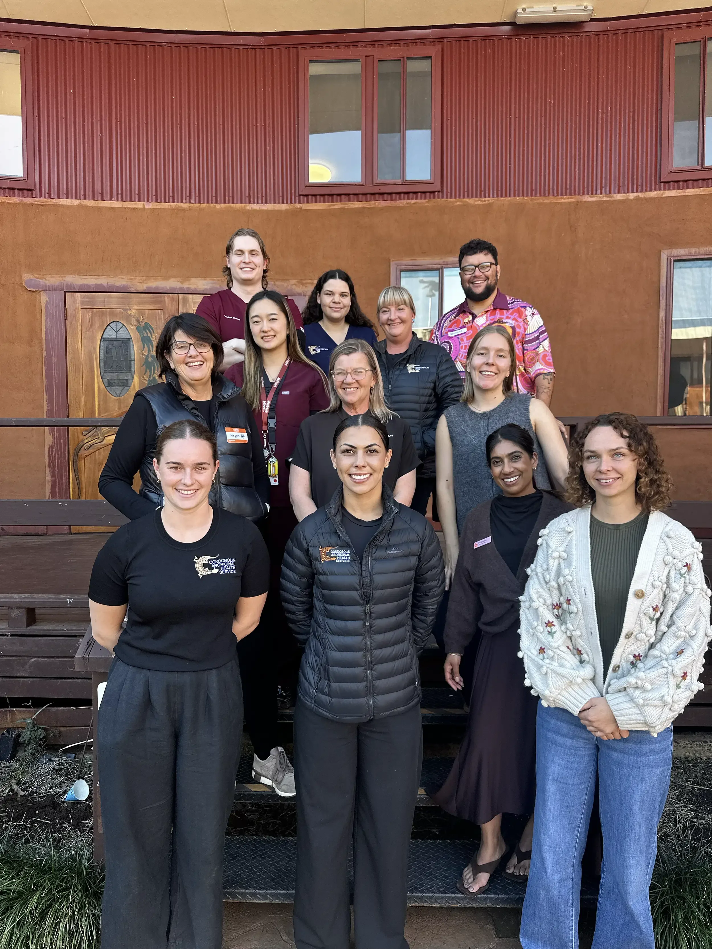 Group of people standing on steps outside a building, smiling, with some wearing name tags and black jackets, in front of a rustic, multi-story building with a red upper wall and brown lower wall.