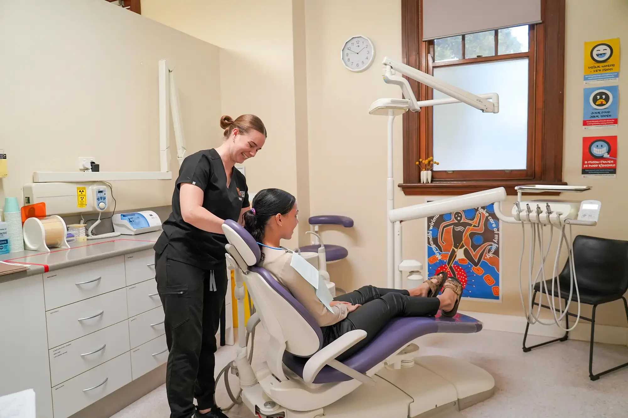 A dental office with a dentist and a patient. The patient is sitting in a dental chair, smiling, while the dentist stands beside her smiling. The room is decorated with colorful artwork and has various dental tools and equipment.