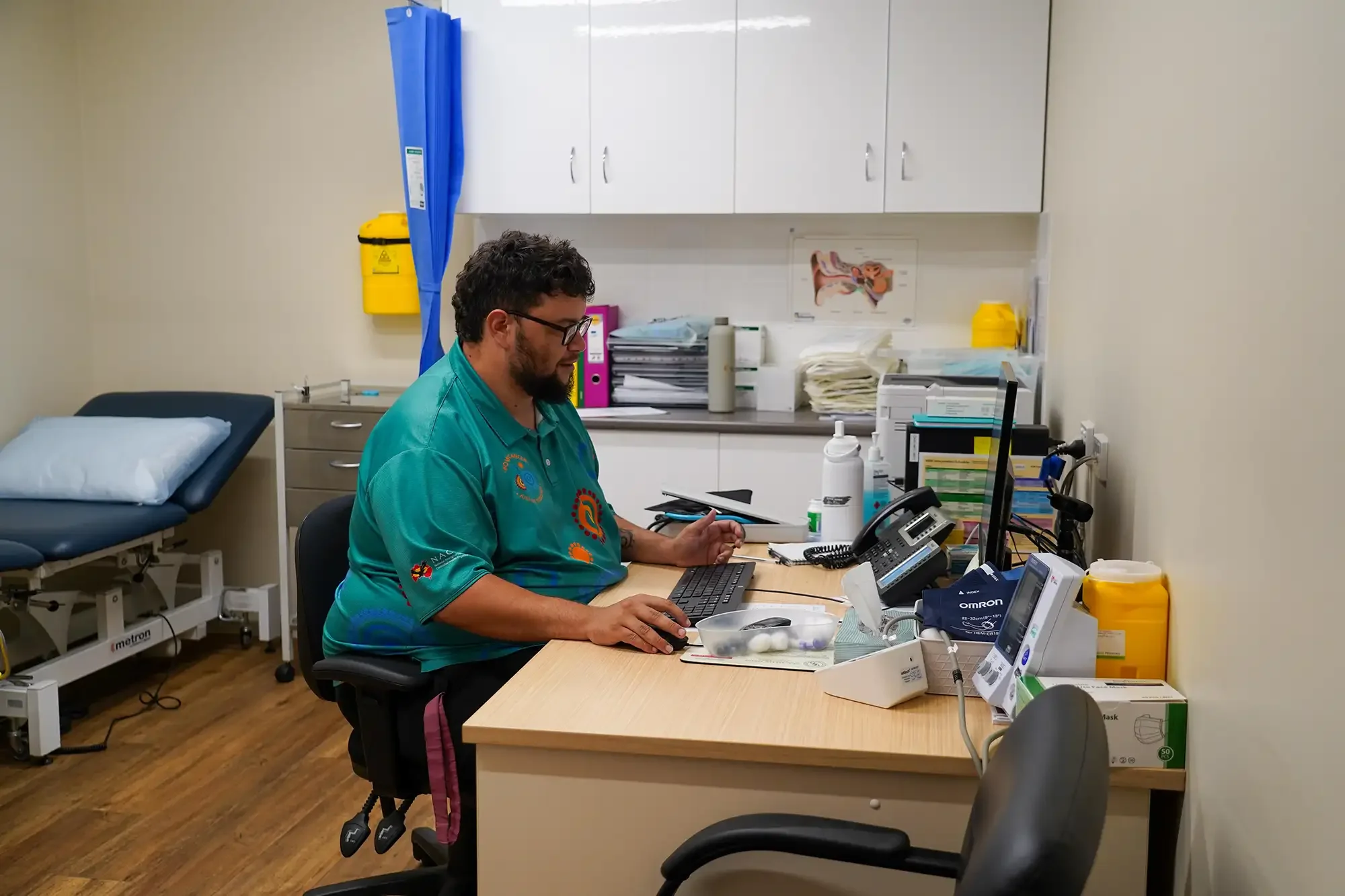 A man wearing glasses and a teal shirt sitting at a desk in a medical office, working on a laptop, with medical supplies and electronic devices on the desk.