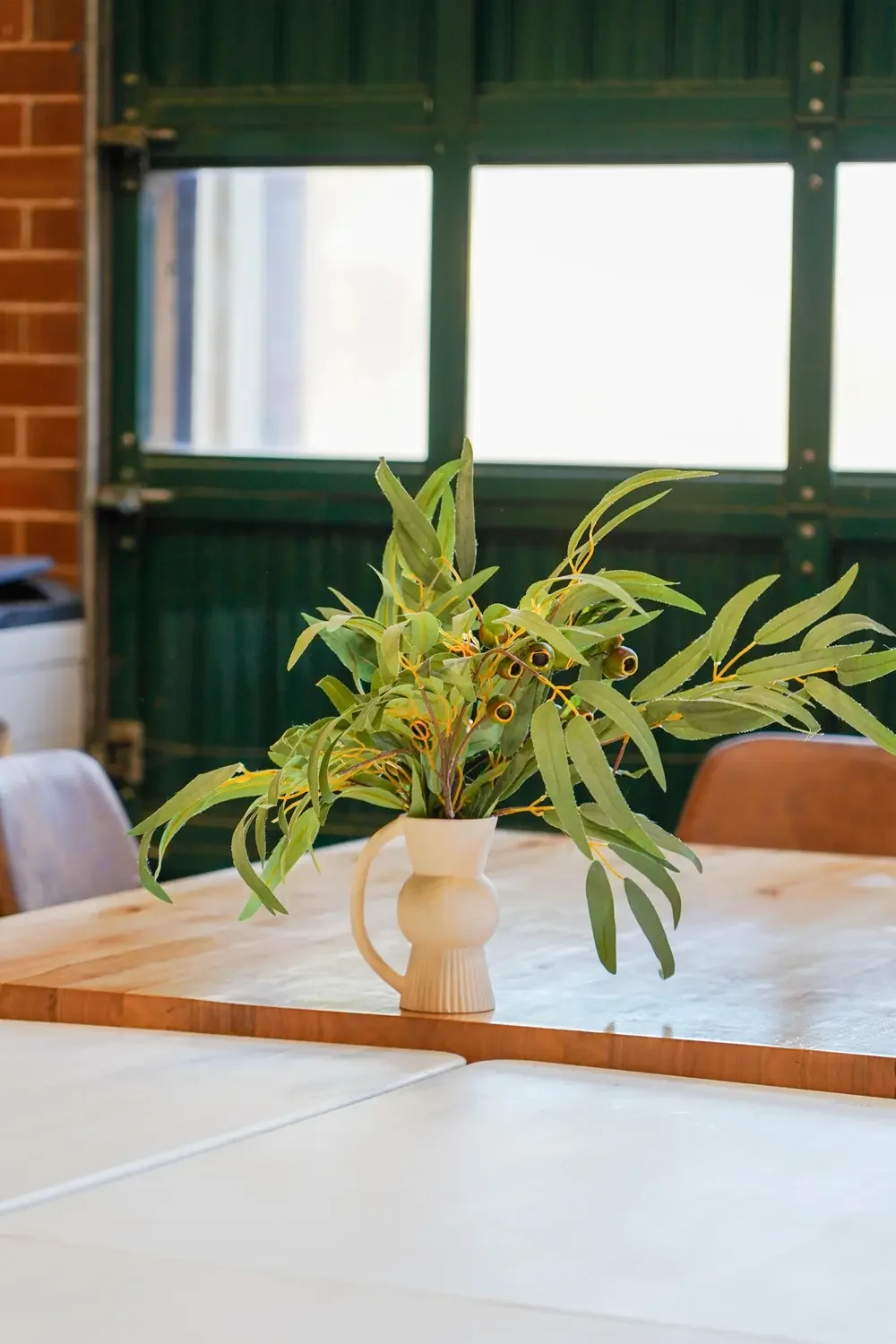 A vase with a green leafy plant on a wooden table inside a room with a green window and brick wall.