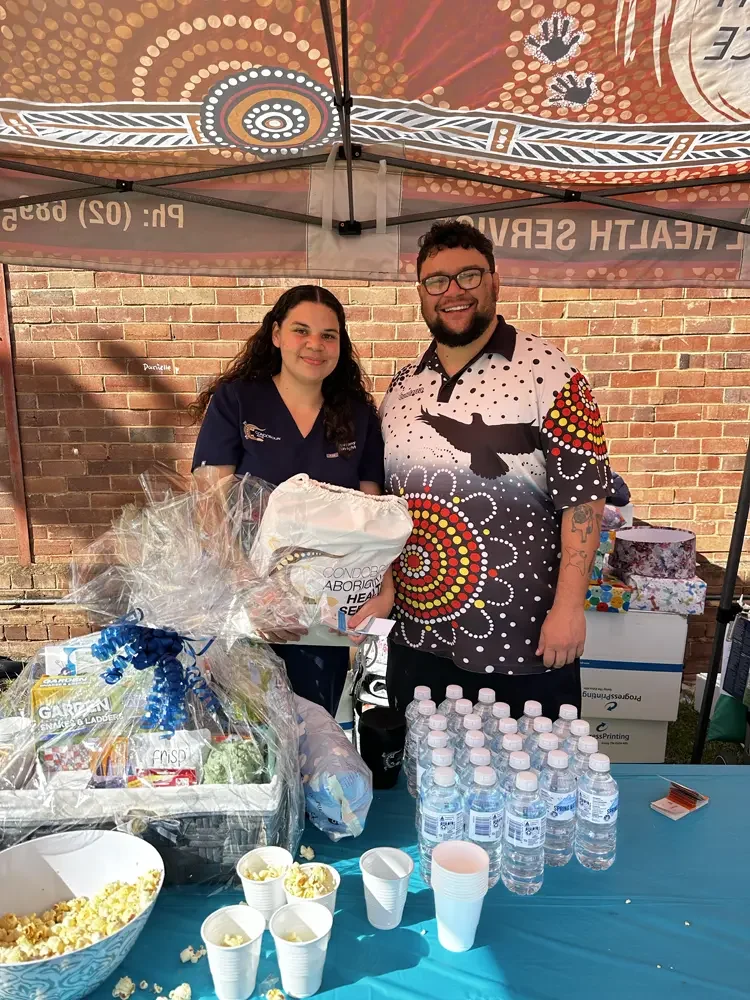 Two people standing behind a table with snacks and water bottles at an outdoor event. They are under a colorful canopy with a brick wall in the background. 