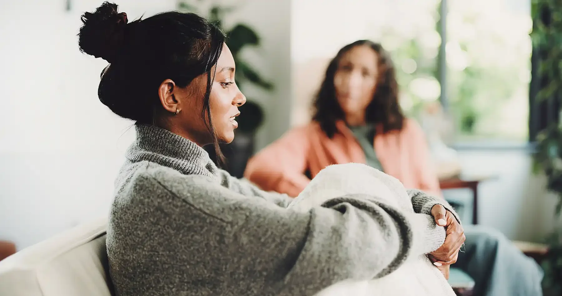 lady sitting on a lounge with her knees to her chest, having a conversation with another lady in the background