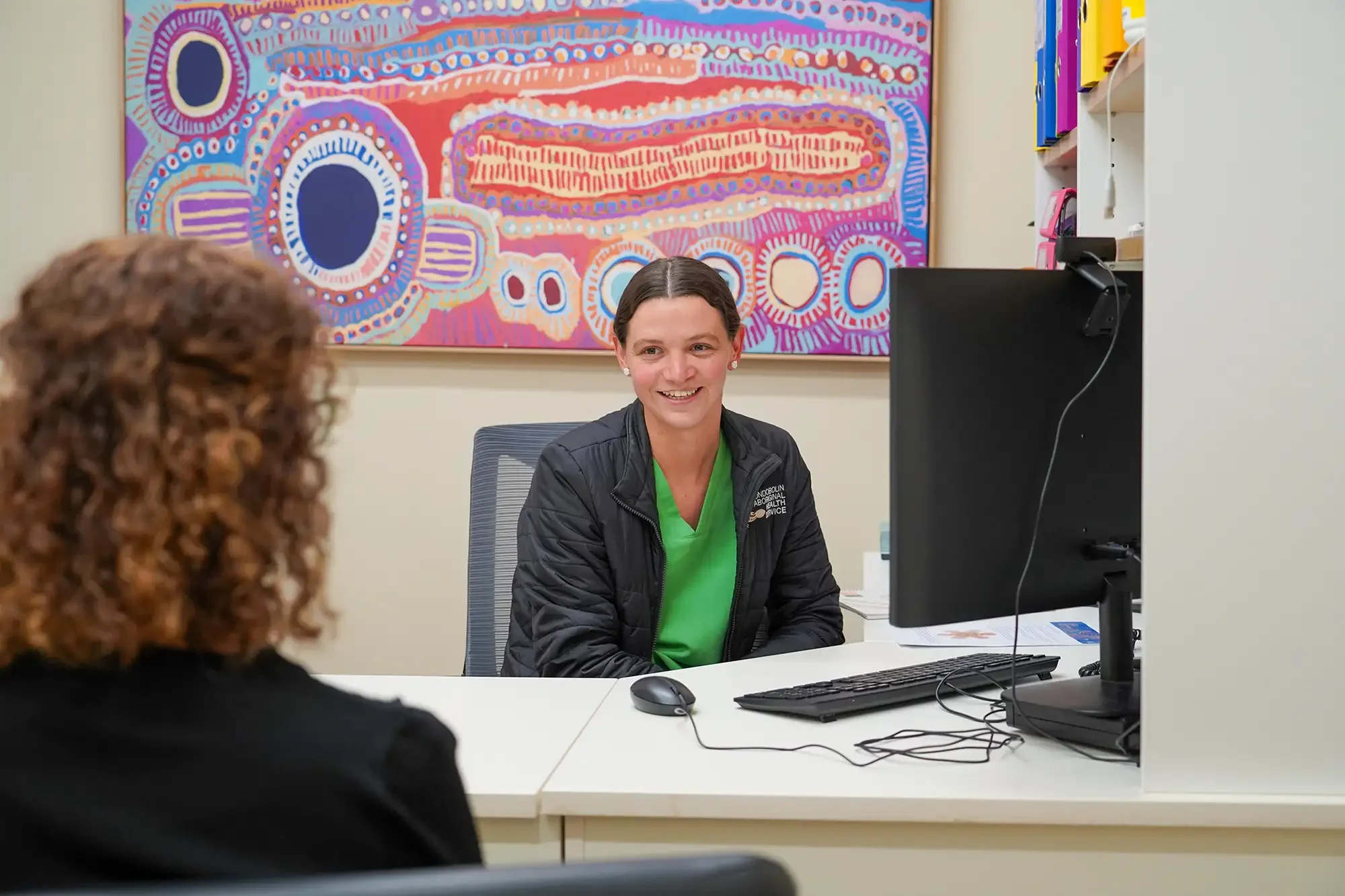 A woman in medical scrubs and a black jacket smiling and talking to a person with curly red hair in a medical or office setting. There is a colorful abstract painting in the background, a computer and desk accessories on the white table.