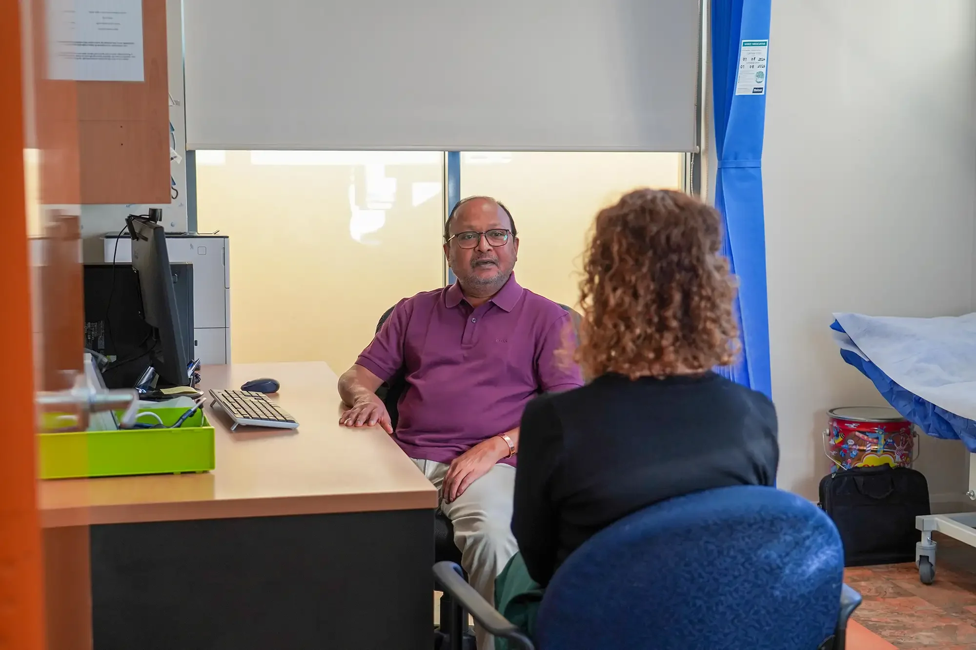 A doctor in a pink polo shirt and glasses is sitting across from a woman with curly hair, wearing a black blazer, in the doctors office. They are engaged in conversation. The room contains medical equipment, a computer, and a bed with blue covers.