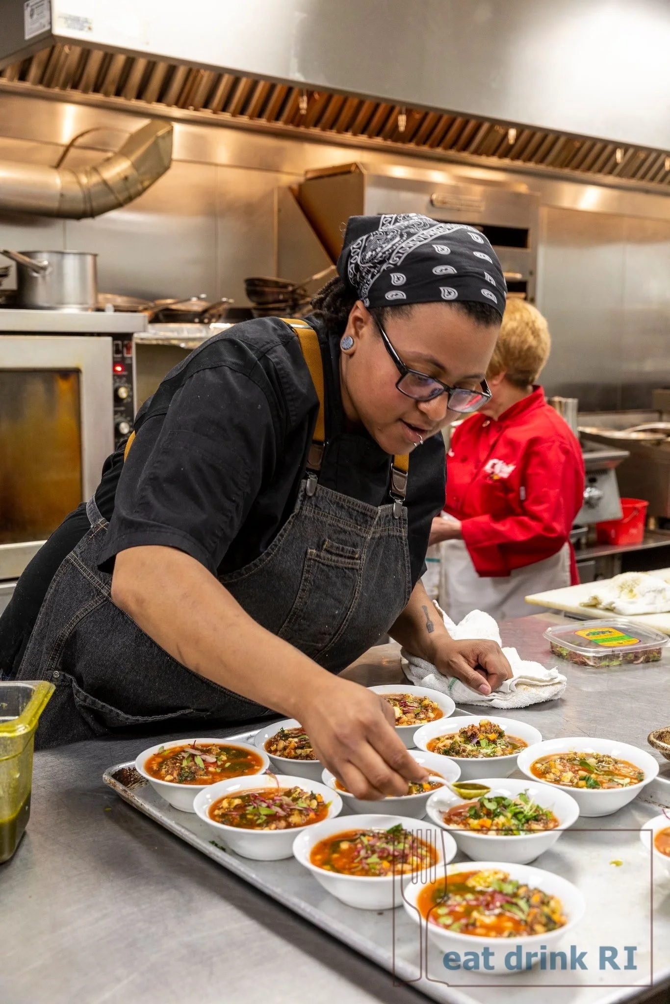 A chef preparing bowls of soup in a commercial kitchen with another person in the background. The bowls contain colorful, garnished soup. The kitchen has stainless steel appliances and a hood vent.