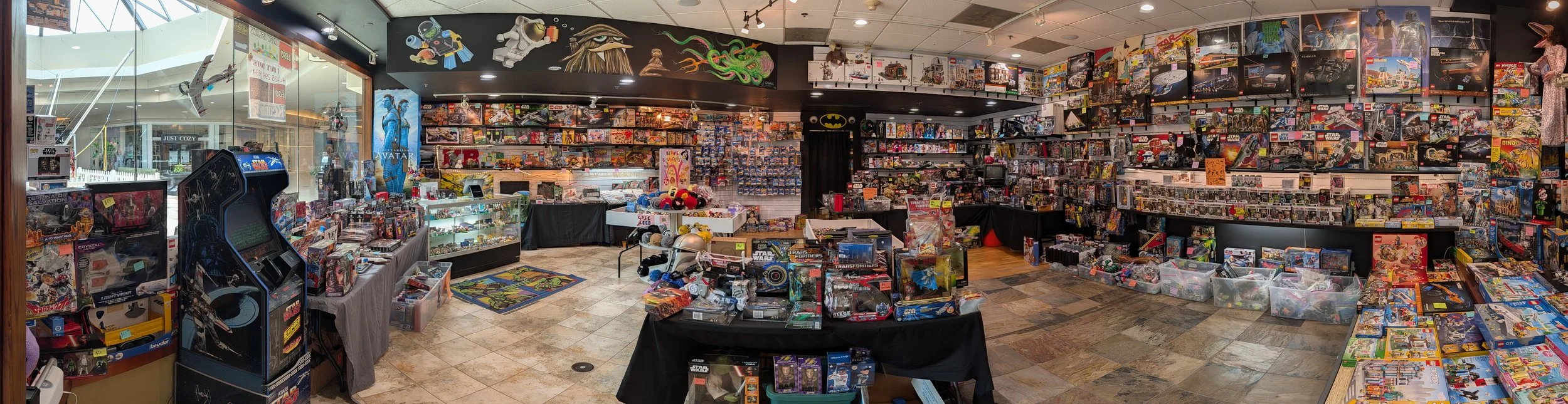 Interior of a toy store filled with shelves and tables displaying toys, action figures, and collectibles, with a window showing an outside shopping mall.