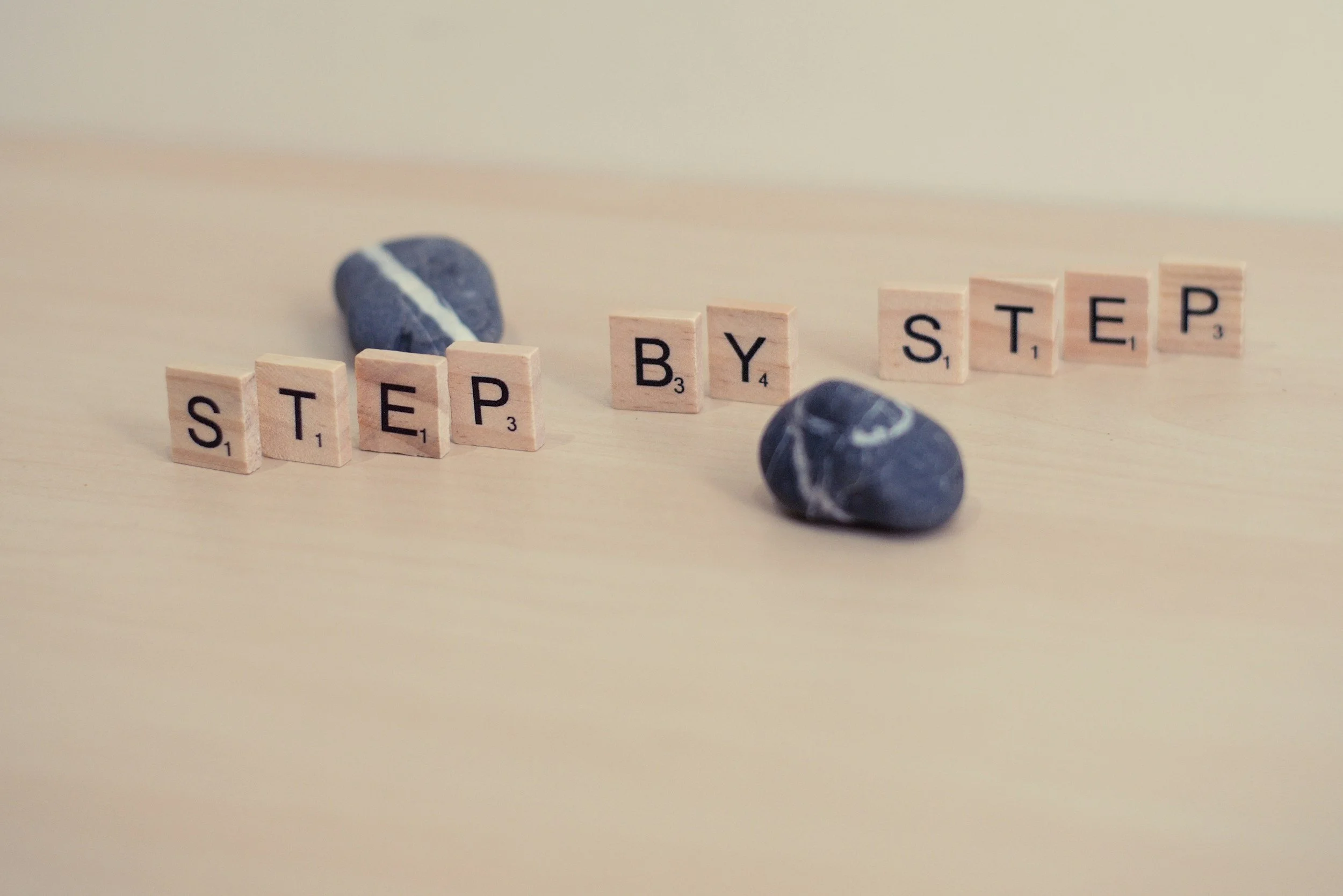 Wooden Scrabble tiles spelling out "STEP BY STEP" on a light-colored surface with two marbled rocks in the background.