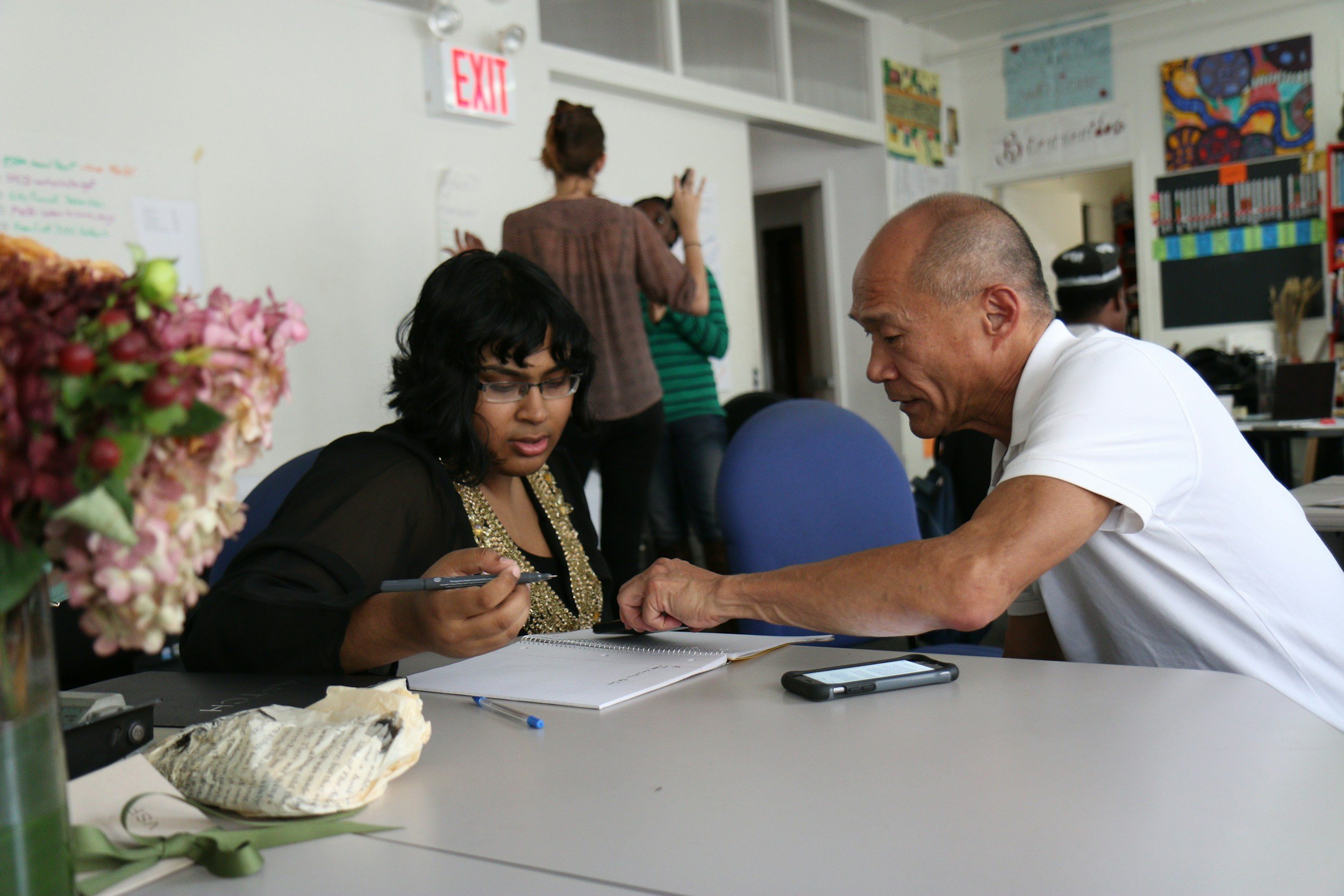 Two people engaged in conversation at a table, with others standing and talking in the background in a classroom or community center setting, decorated with colorful artwork and an EXIT sign on the wall.