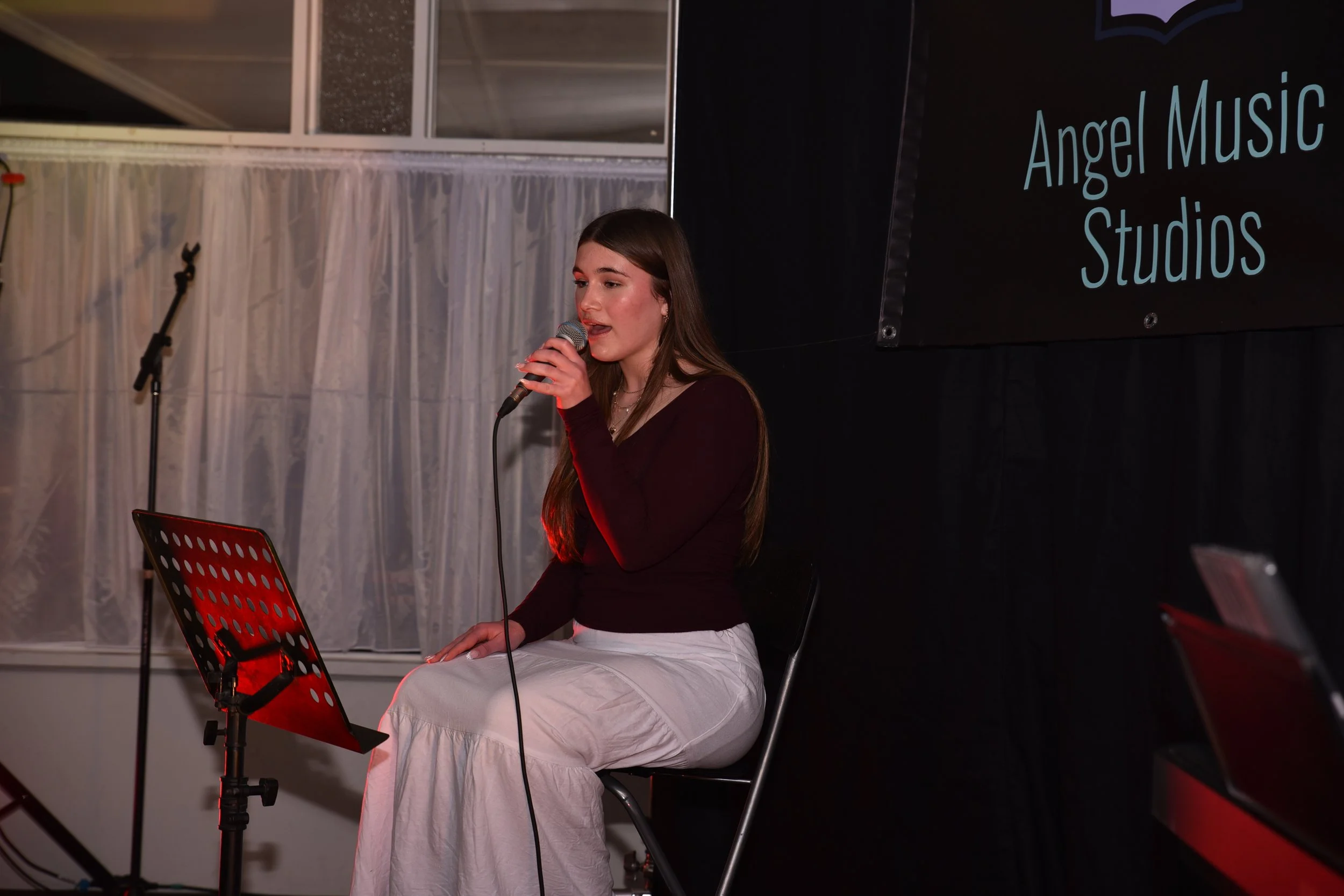A young woman sitting and singing into a microphone on stage during a performance at Angel Music Studios, with a black backdrop and music stand in front of her.
