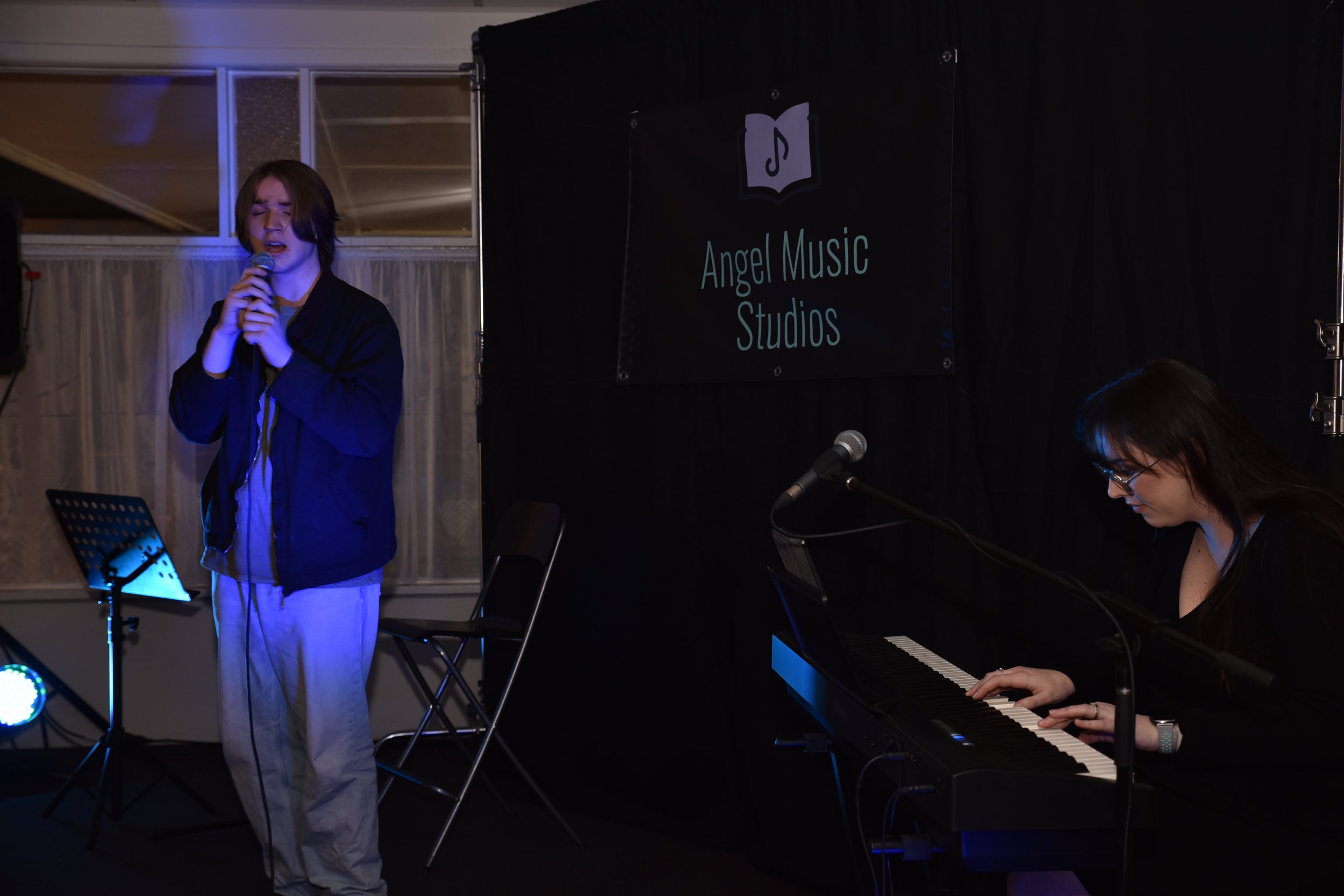 A young woman with short brown hair and a black jacket is singing into a microphone on a stage. A woman with glasses and dark hair is playing a keyboard nearby. The stage has a sign that reads 'Angel Music Studios' with a music book and note logo. Th