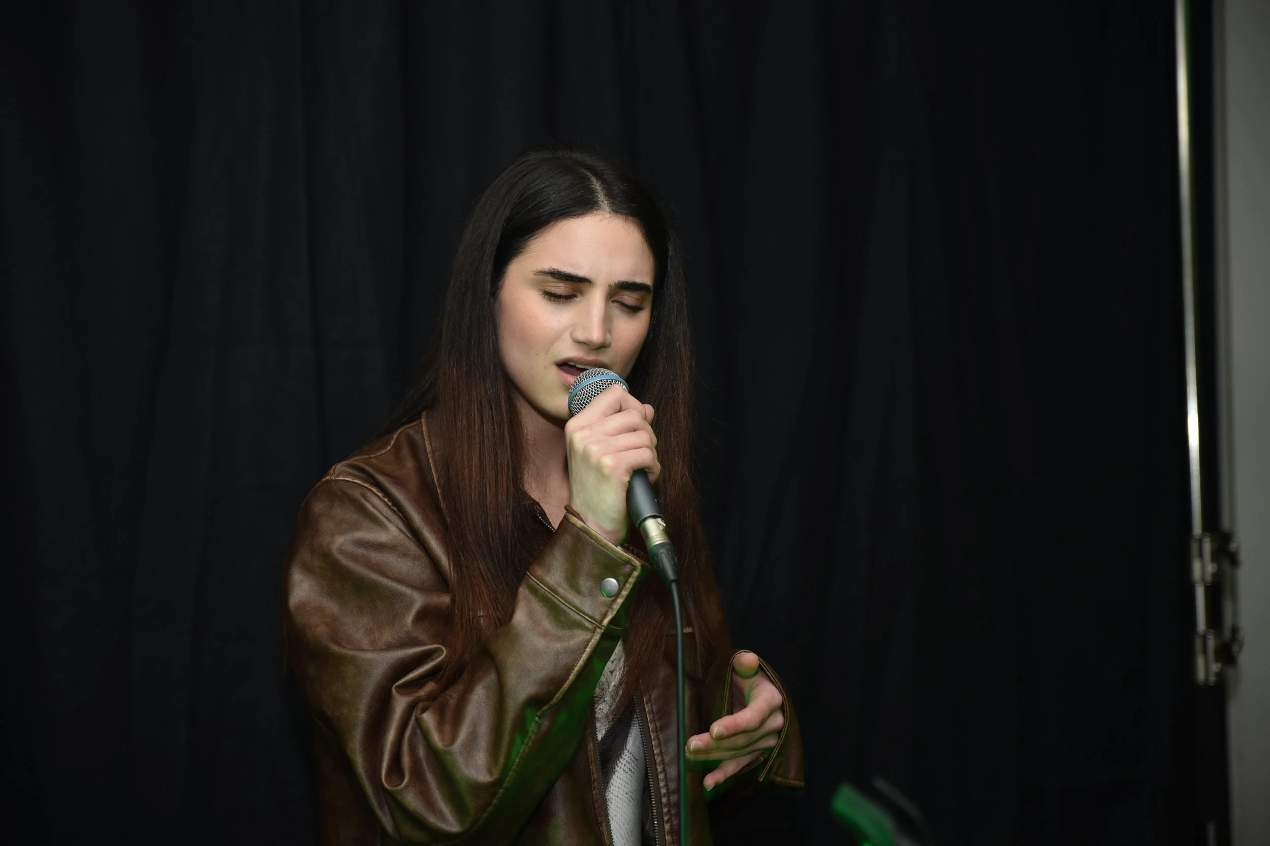 A young woman with long dark hair and fair skin sings into a microphone against a black background, wearing a brown leather jacket.