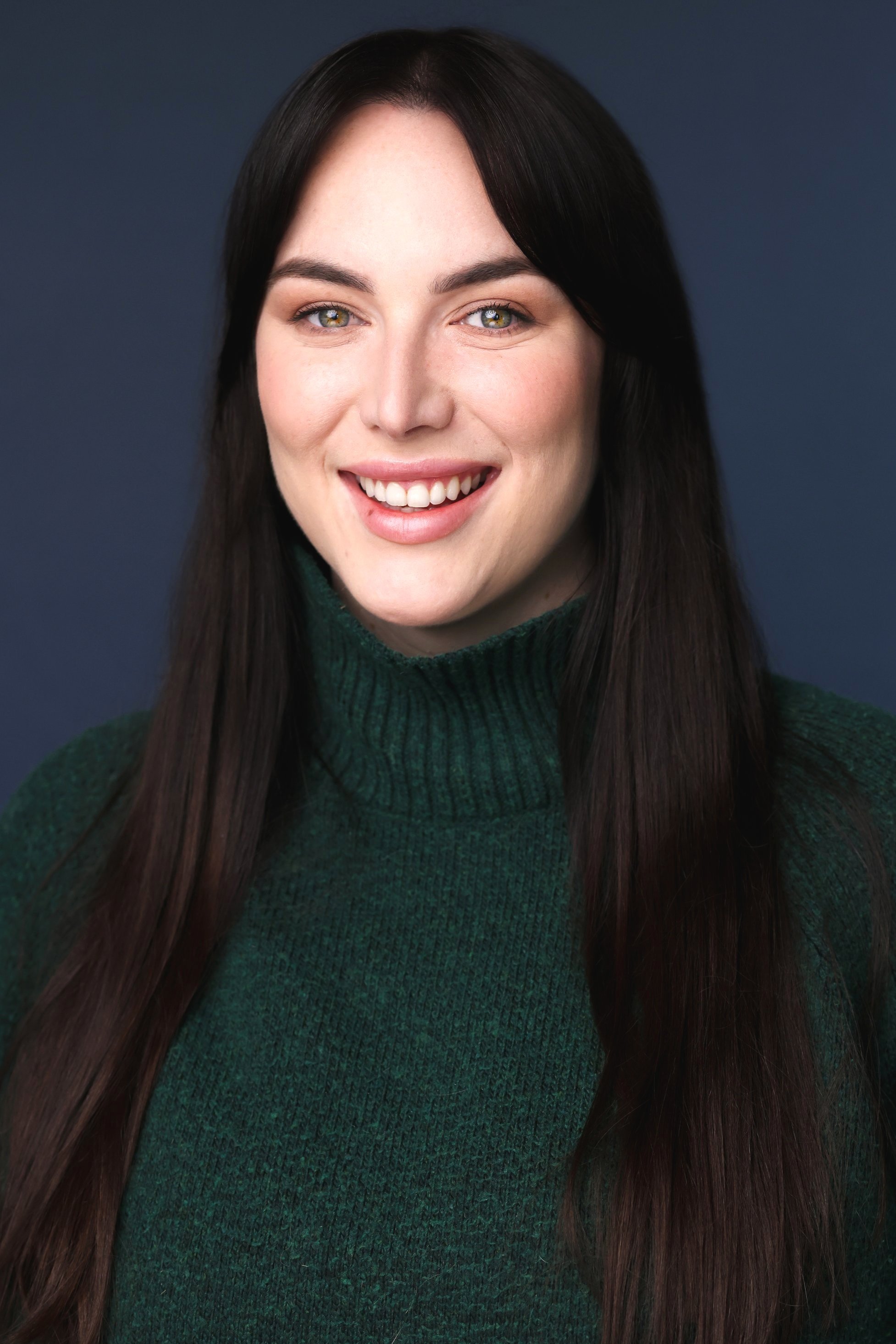 Headshot of a woman with long dark hair, smiling, wearing a dark green turtleneck, set against a dark background.