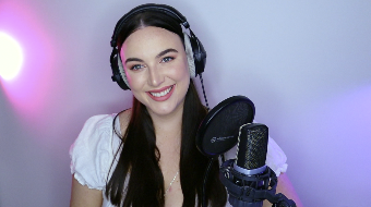 A woman with long dark hair wearing headphones, smiling, and speaking into a microphone in a recording studio with purple and pink lighting.