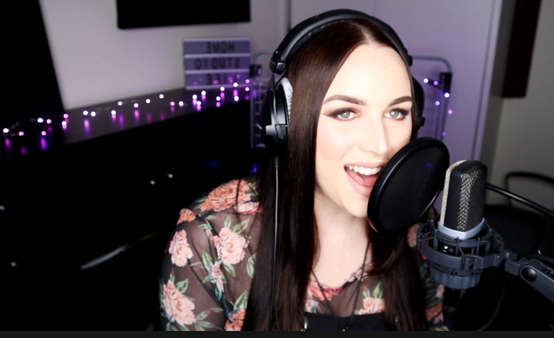 A woman with long brown hair and makeup, wearing headphones and a floral top, is speaking or singing into a microphone in a recording studio.