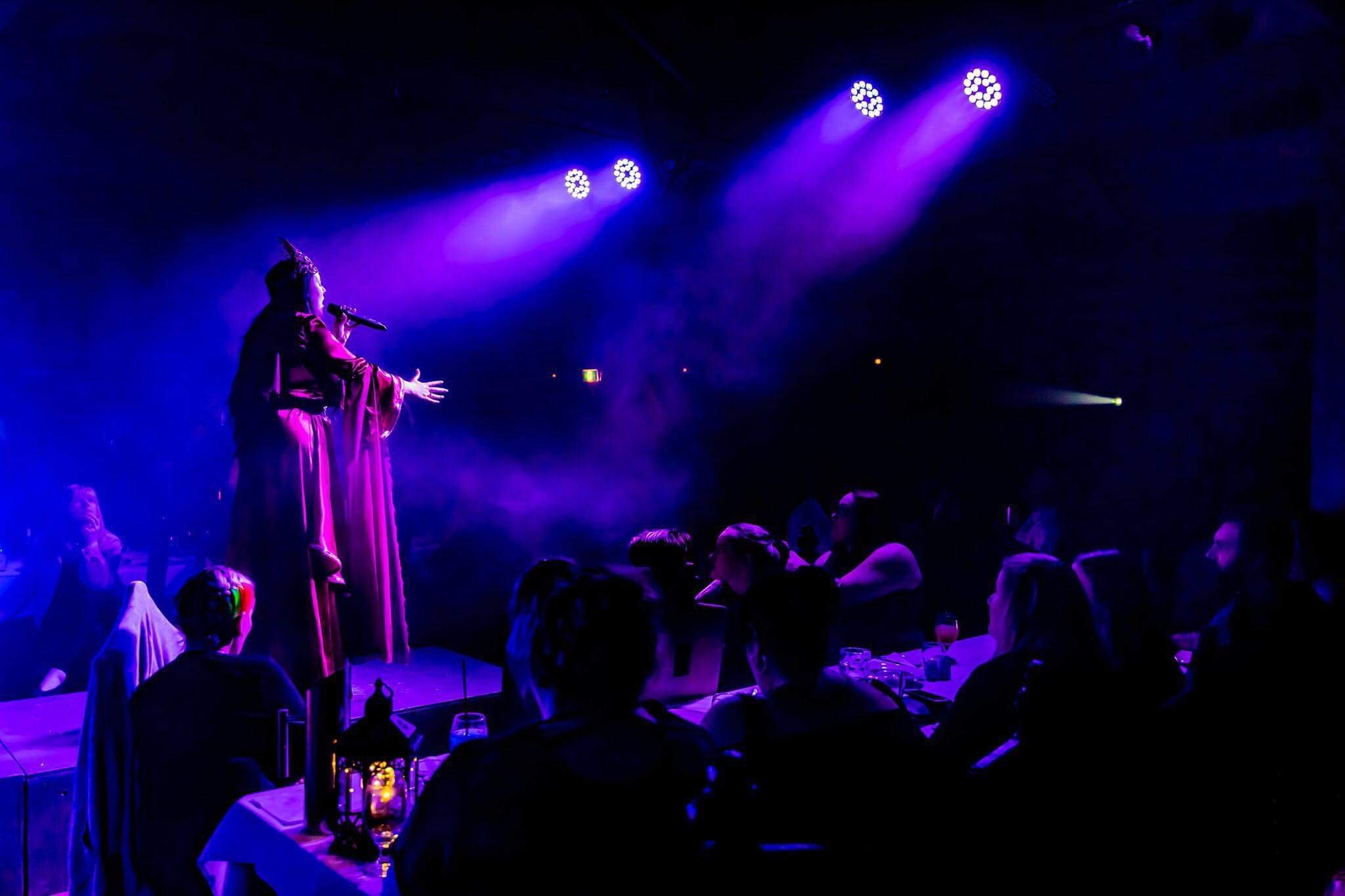 A performer on stage singing into a microphone with purple stage lights shining down. Audience members are seated at tables in a darkened room.