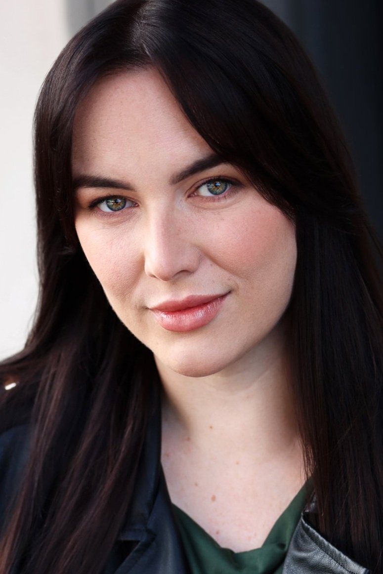Close-up portrait of a woman with long dark hair, blue eyes, light skin, and a subtle smile, wearing a dark jacket.