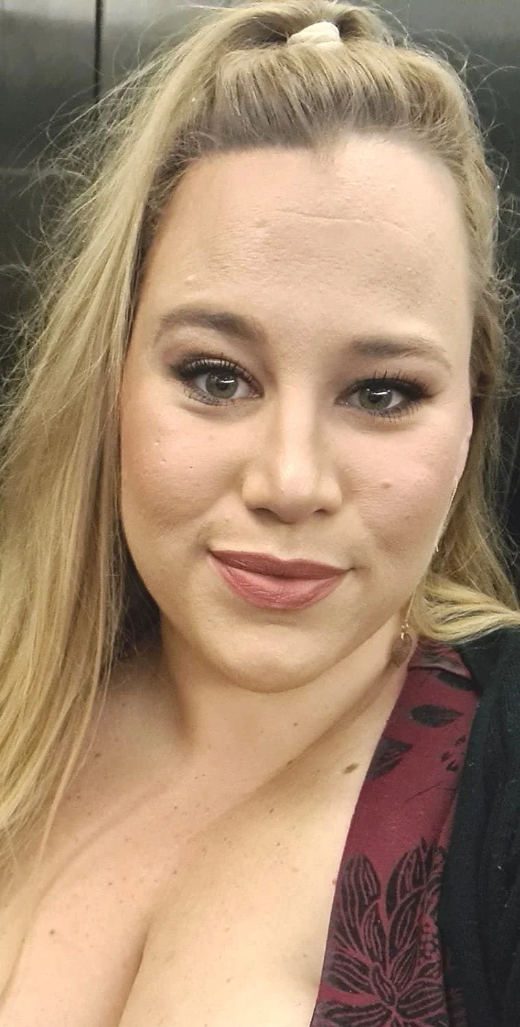 Close-up selfie of a woman with blonde hair, green eyes, wearing makeup, and a maroon and black patterned top.