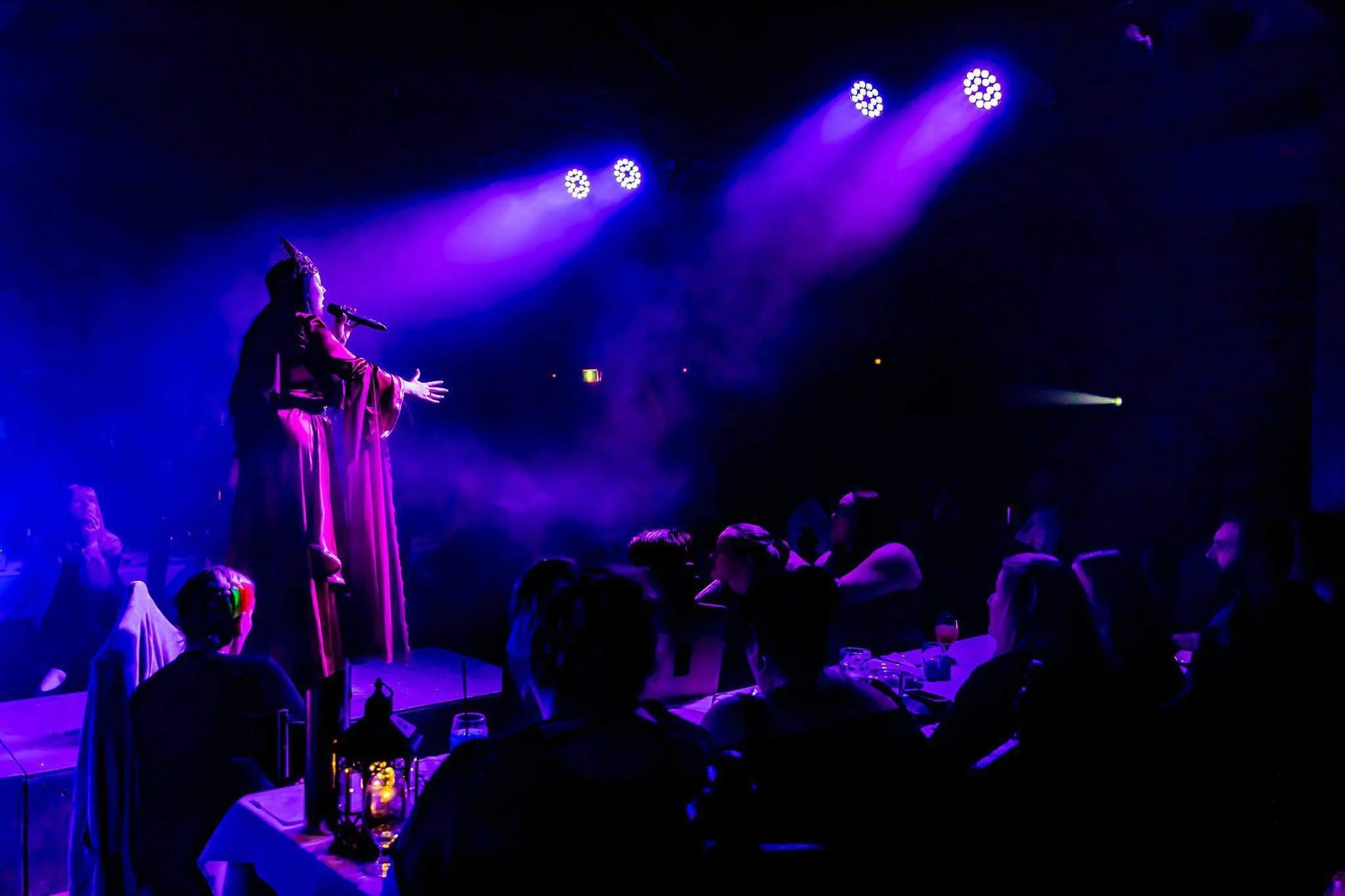 Performer singing on stage with purple lighting, audience seated at tables in a dimly lit venue.