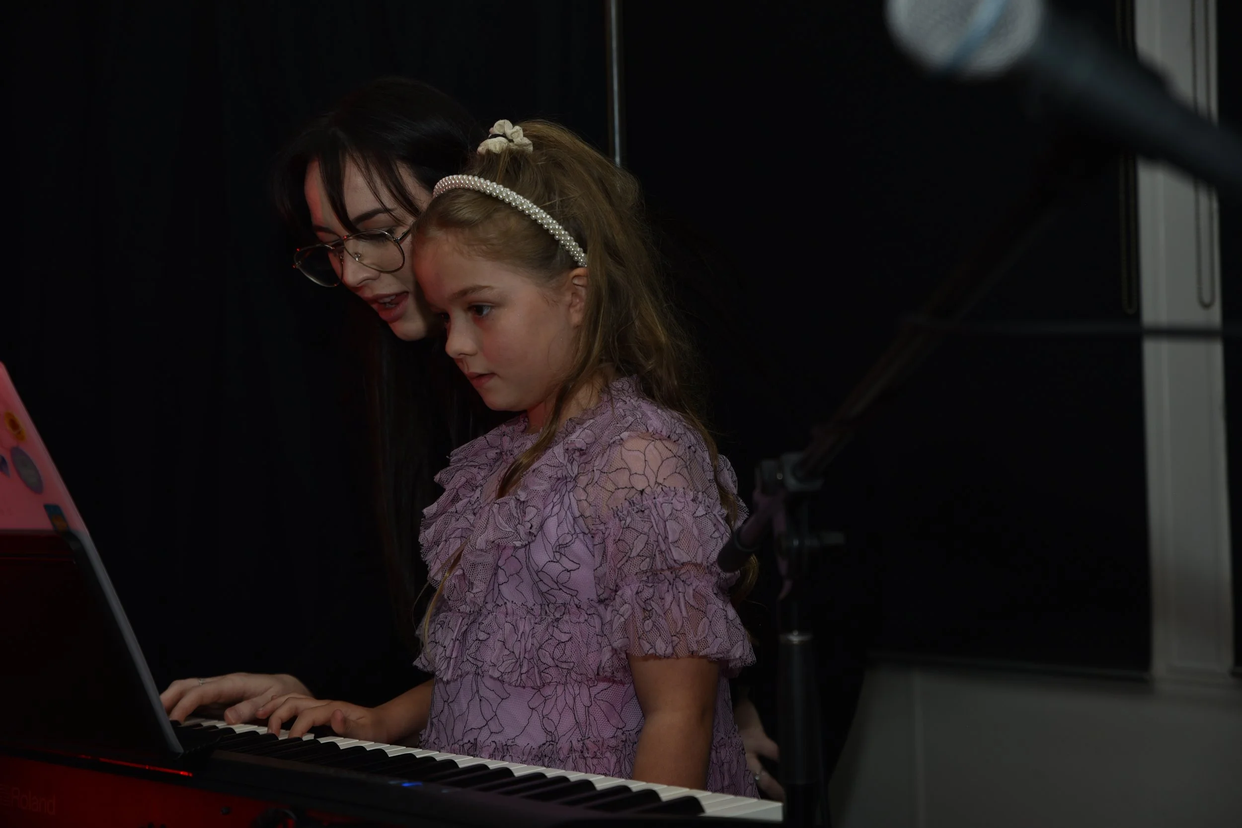 A woman with dark hair, glasses, and a black shirt playing a red piano while a young girl with long hair, a pearl headband, and a pink lace dress watches.